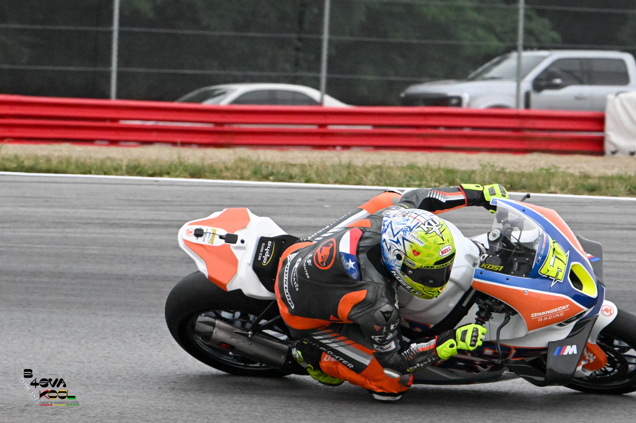 A motorcycle racer in full gear leaning into a turn on a racetrack, with a blurred background of cars and fencing.