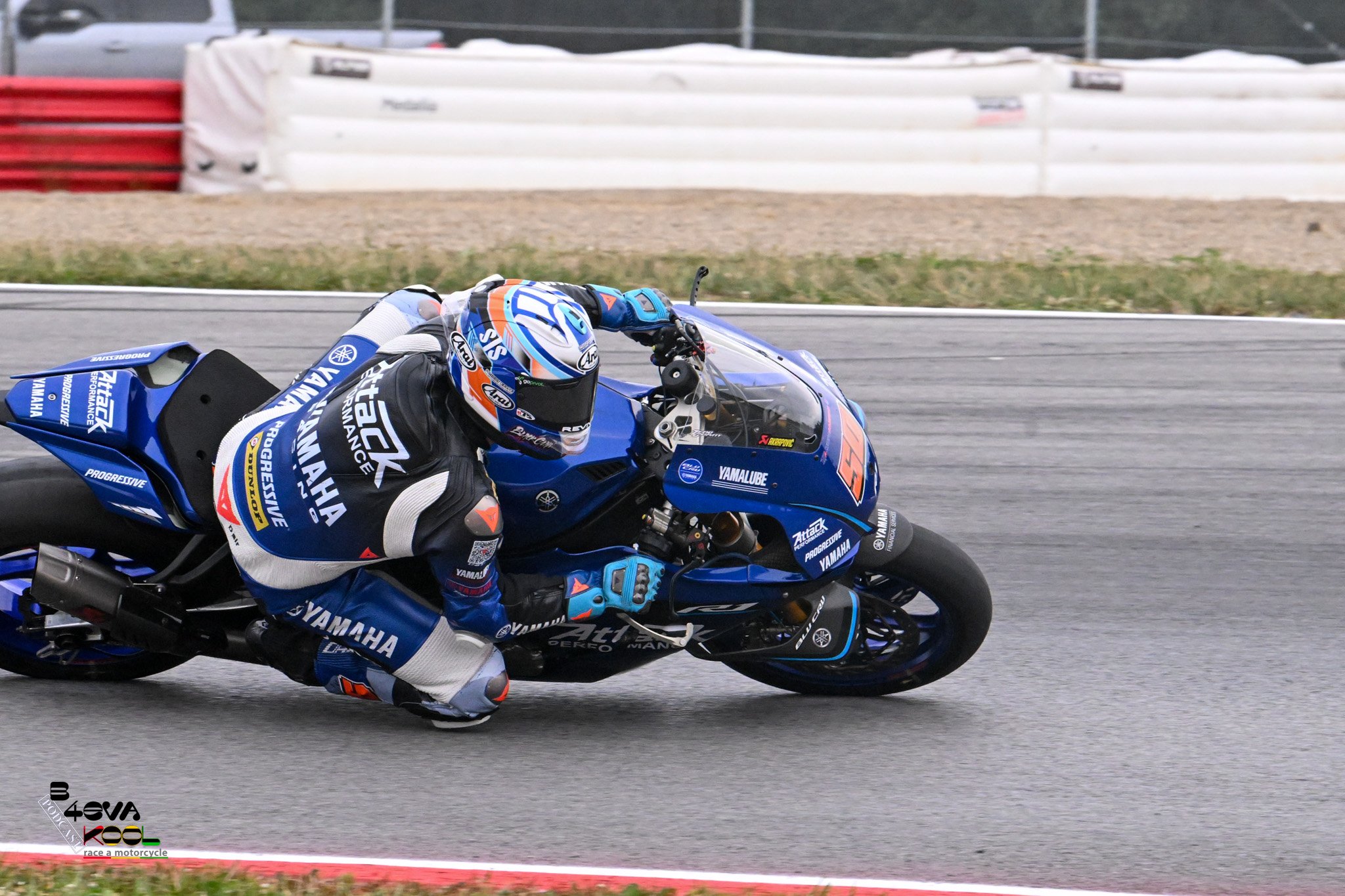 Motorcycle racer leaning into a turn on a racetrack, wearing a blue, white, and black racing suit and helmet with Yamaha logos, on a blue Yamaha motorcycle.