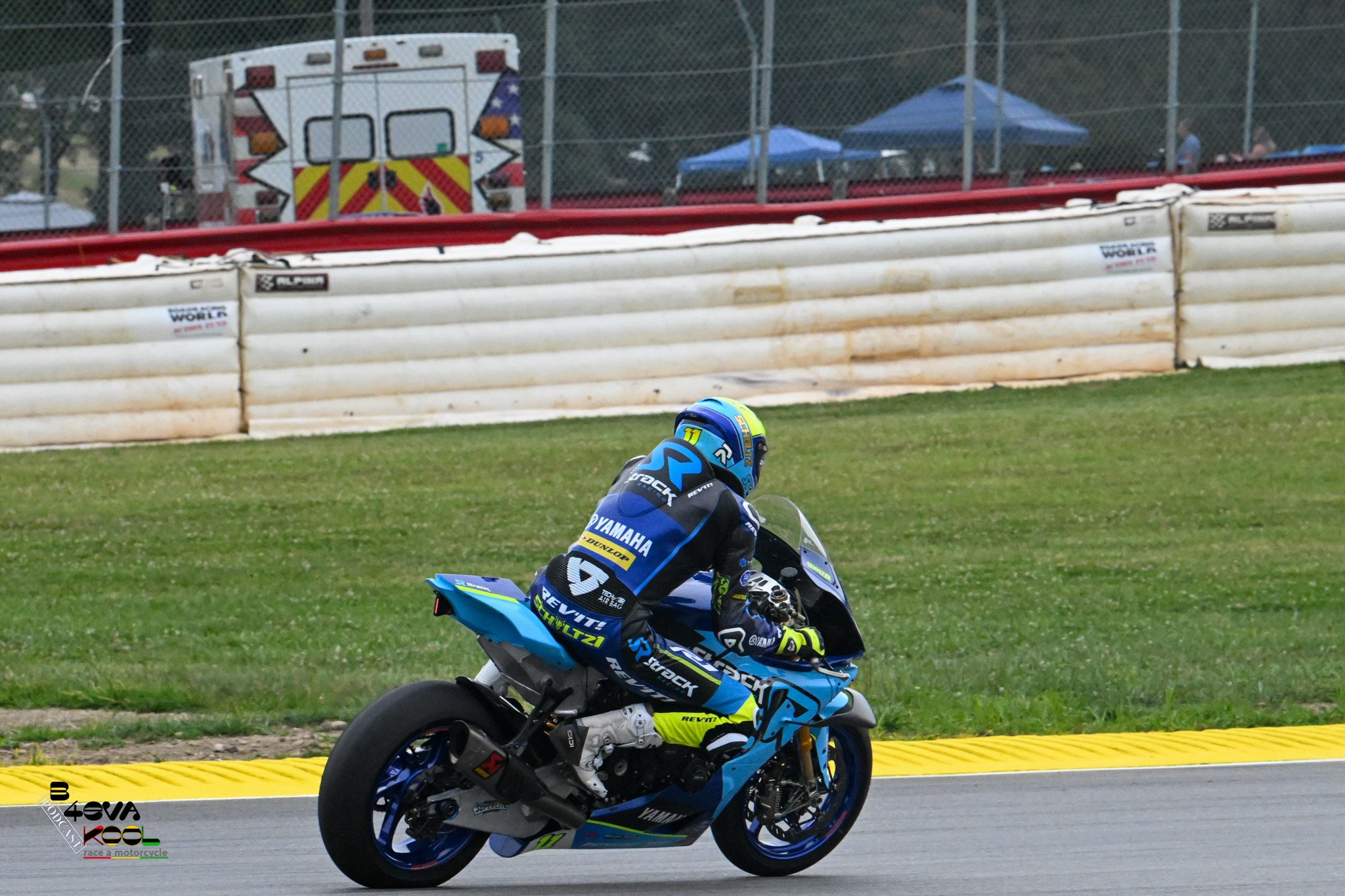 A motorcycle racer in blue and black gear riding a blue Yamaha motorcycle on a racetrack with a barrier and emergency vehicle in the background.