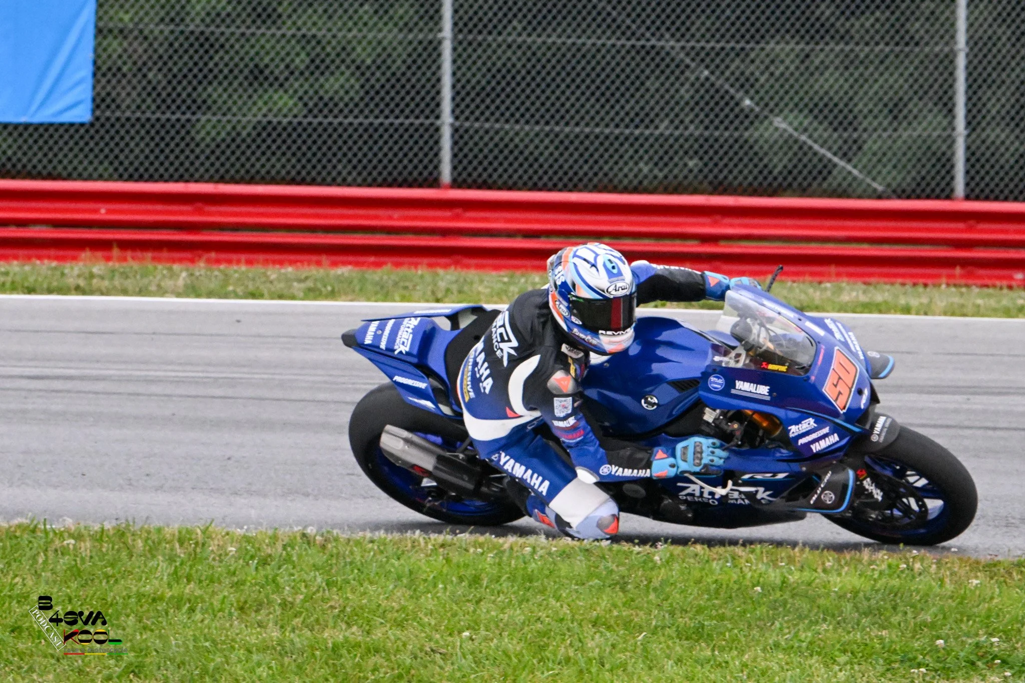 Motorcycle racer leaning into a turn on a race track, dressed in a blue racing suit and helmet, riding a blue Yamaha motorcycle with various sponsor logos, with a fence and greenery in the background.