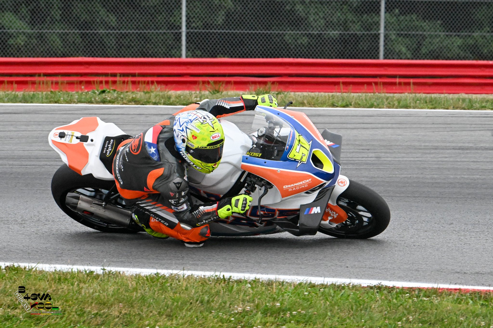 Motorcycle racer wearing a yellow helmet and colorful suit leaning into a turn on a racetrack with red barriers and green grass in the background.