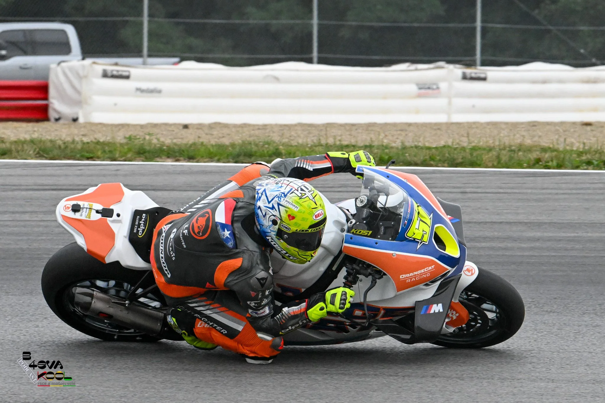 Motorcycle racer leaning into a turn on a race track, dressed in orange, black, and yellow gear, with a blue, white, and orange motorcycle.