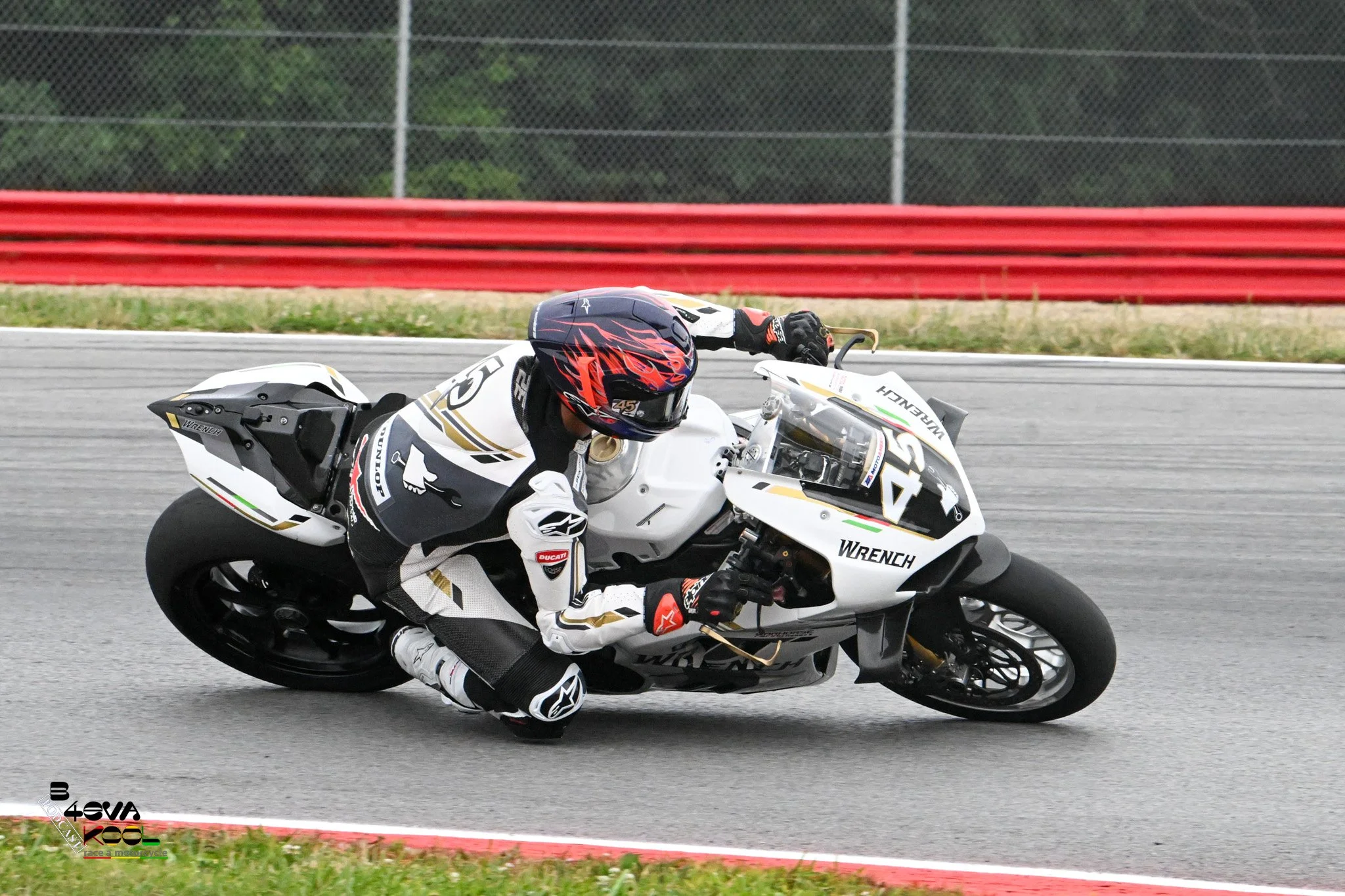 Motorcycle racer leaning into a turn on a racetrack, wearing a white racing suit and a black helmet with red flames.