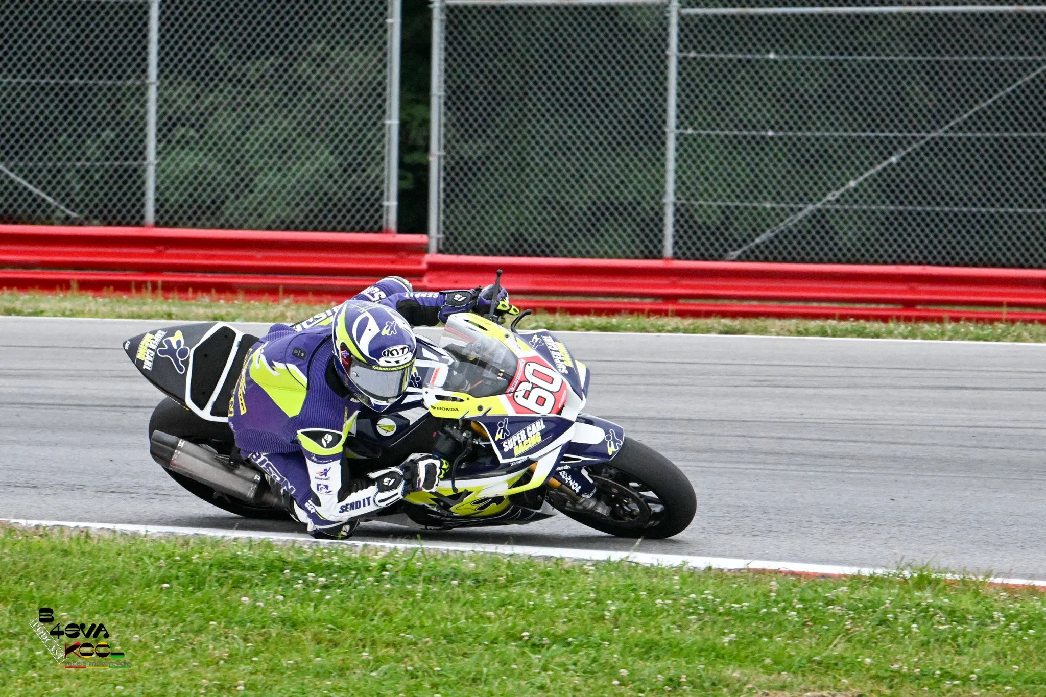 Motorcycle racer leaning into a turn on a race track with a chain-link fence in the background.