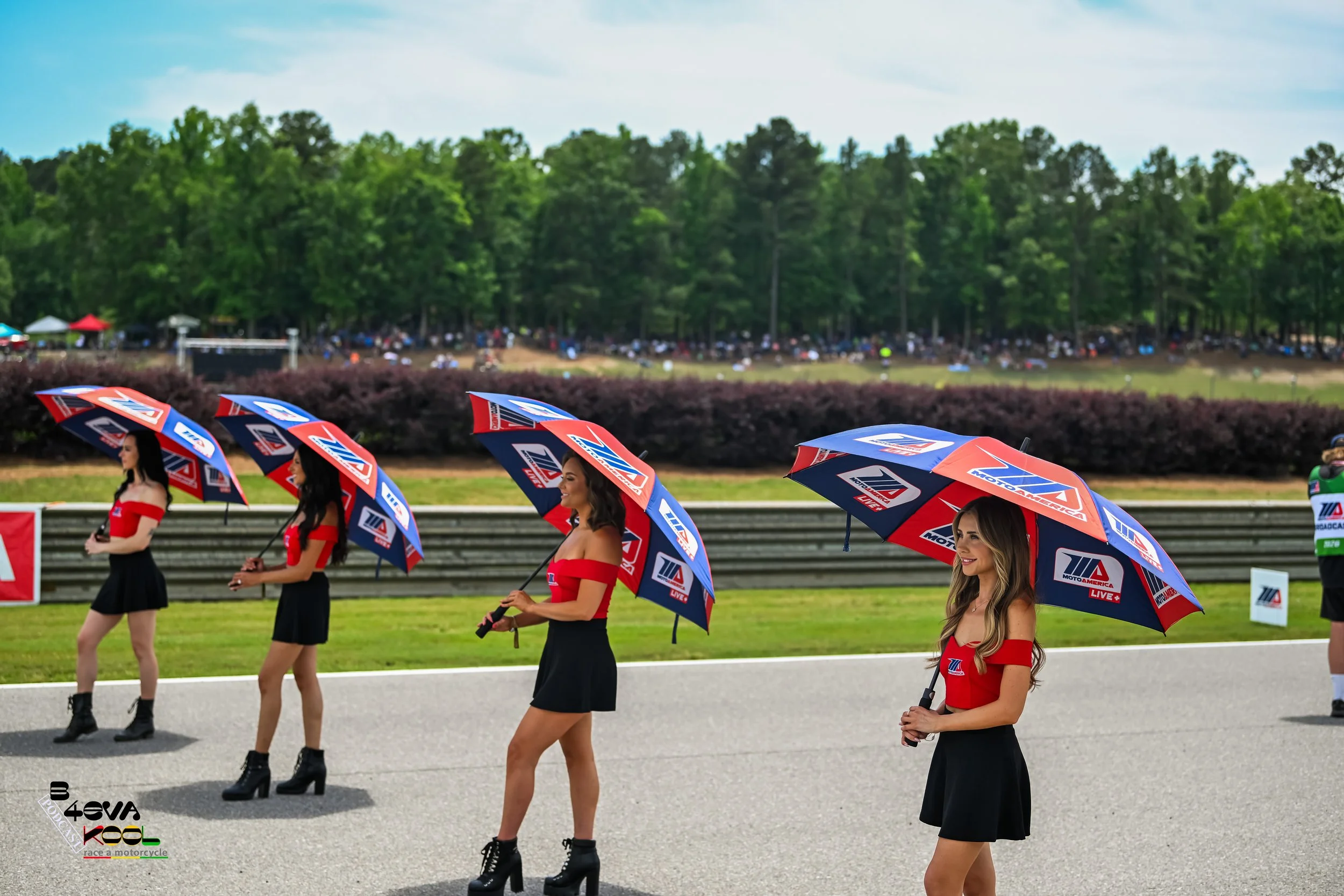 Five women in red tops and black skirts holding umbrellas with motorsport logos standing in a row at a racing event. Spectators in the background sit on a wooded hillside.