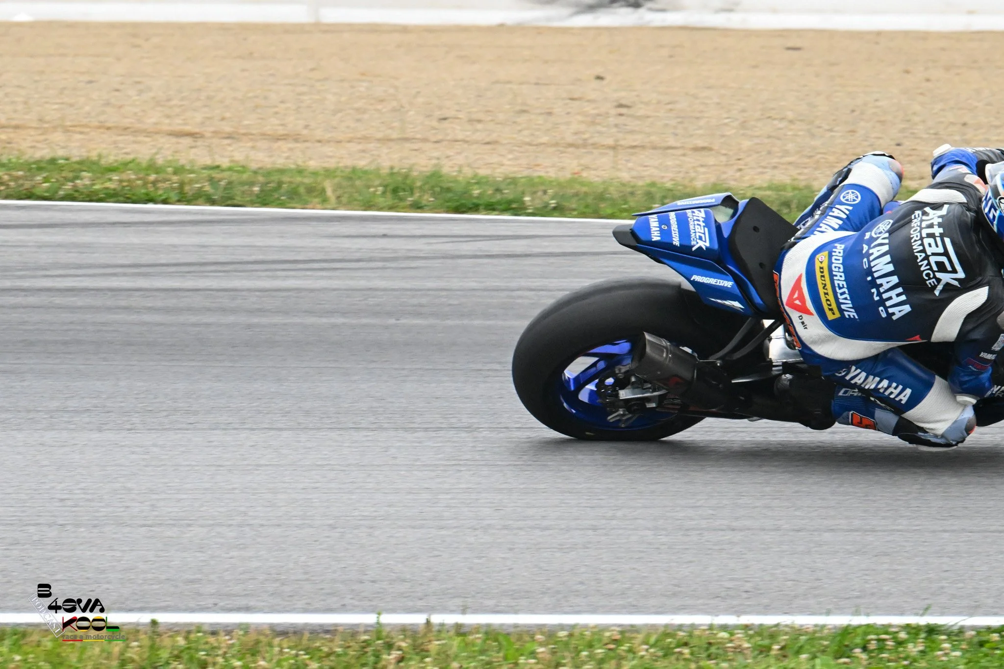 A motorcycle racer leaning into a turn on a racetrack, wearing a blue and black racing suit and helmet, with sponsorship logos visible.