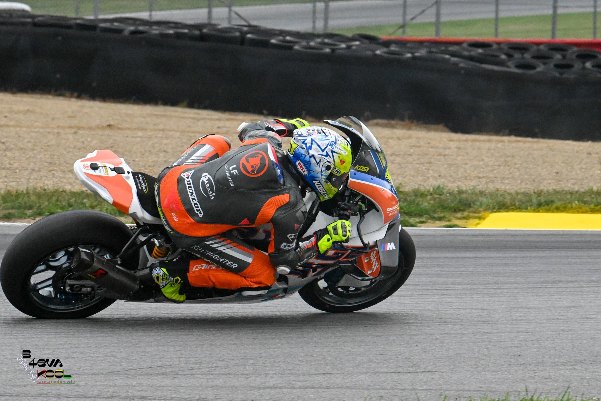 Motorcycle racer in an orange and black racing suit leaning into a turn on a race track, with a black safety barrier and green grass in the background.