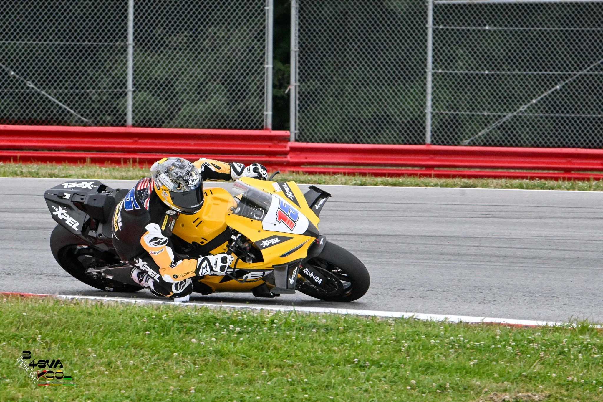 Motorcycle racer on a yellow and black bike speeding on a race track with red safety barriers and a chain-link fence in the background.
