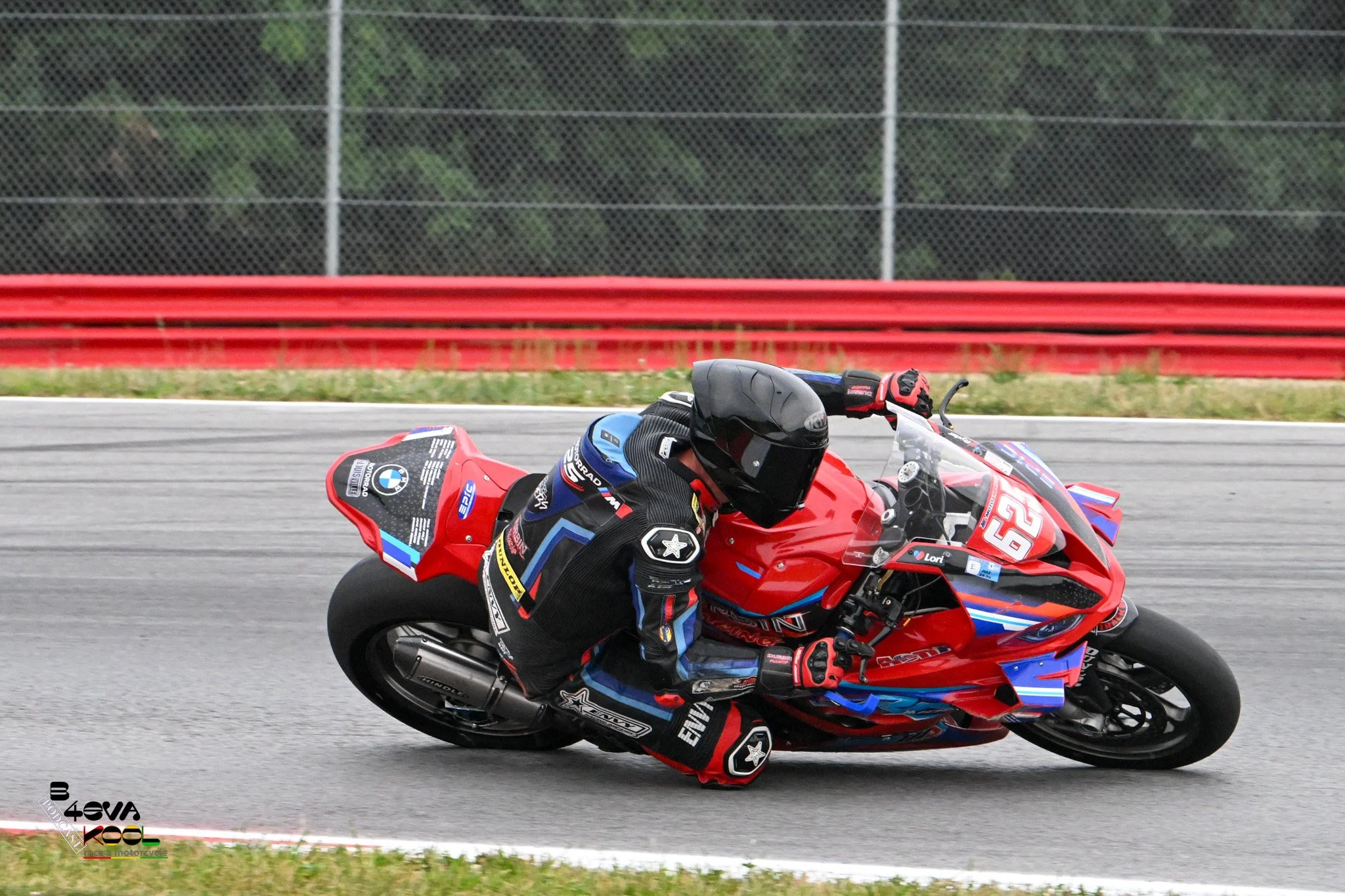 Motorcycle racer leaning into a turn on a race track, wearing a racing suit and helmet, with a red and blue motorcycle.
