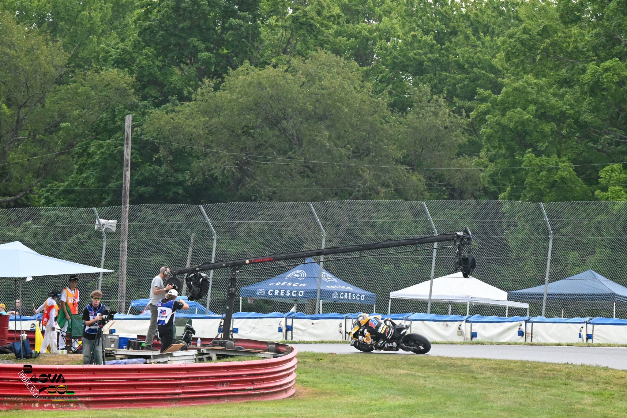 Professional motorcycle race at a track with crew members, tents, and safety barriers, with green trees in the background.