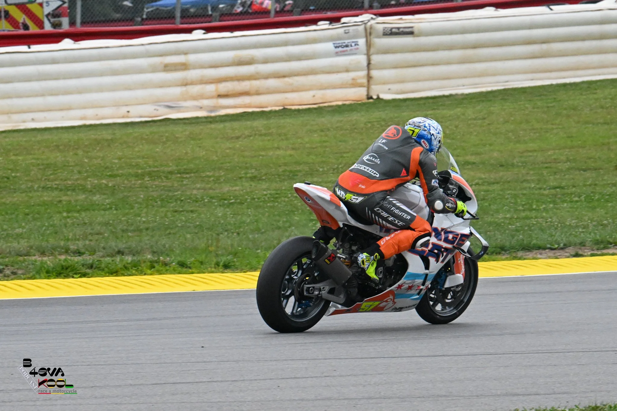 A motorcycle racer in a racing suit and helmet riding a racing motorcycle on a track with green grass and a safety barrier in the background.