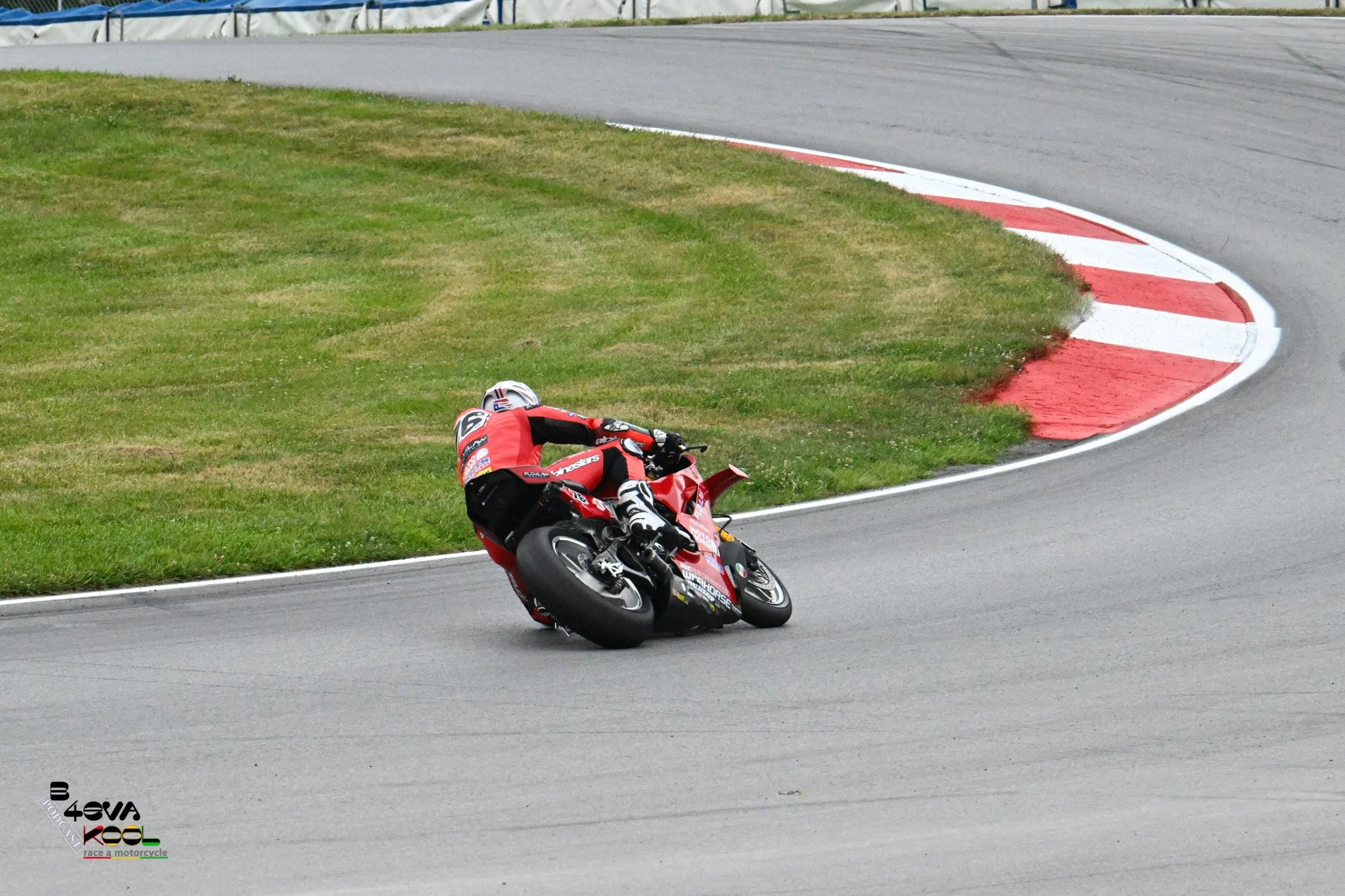 A race motorcycle rider in a red racing suit and helmet falling off a red motorcycle on a curved section of a racetrack, with green grass and a red and white curb nearby.