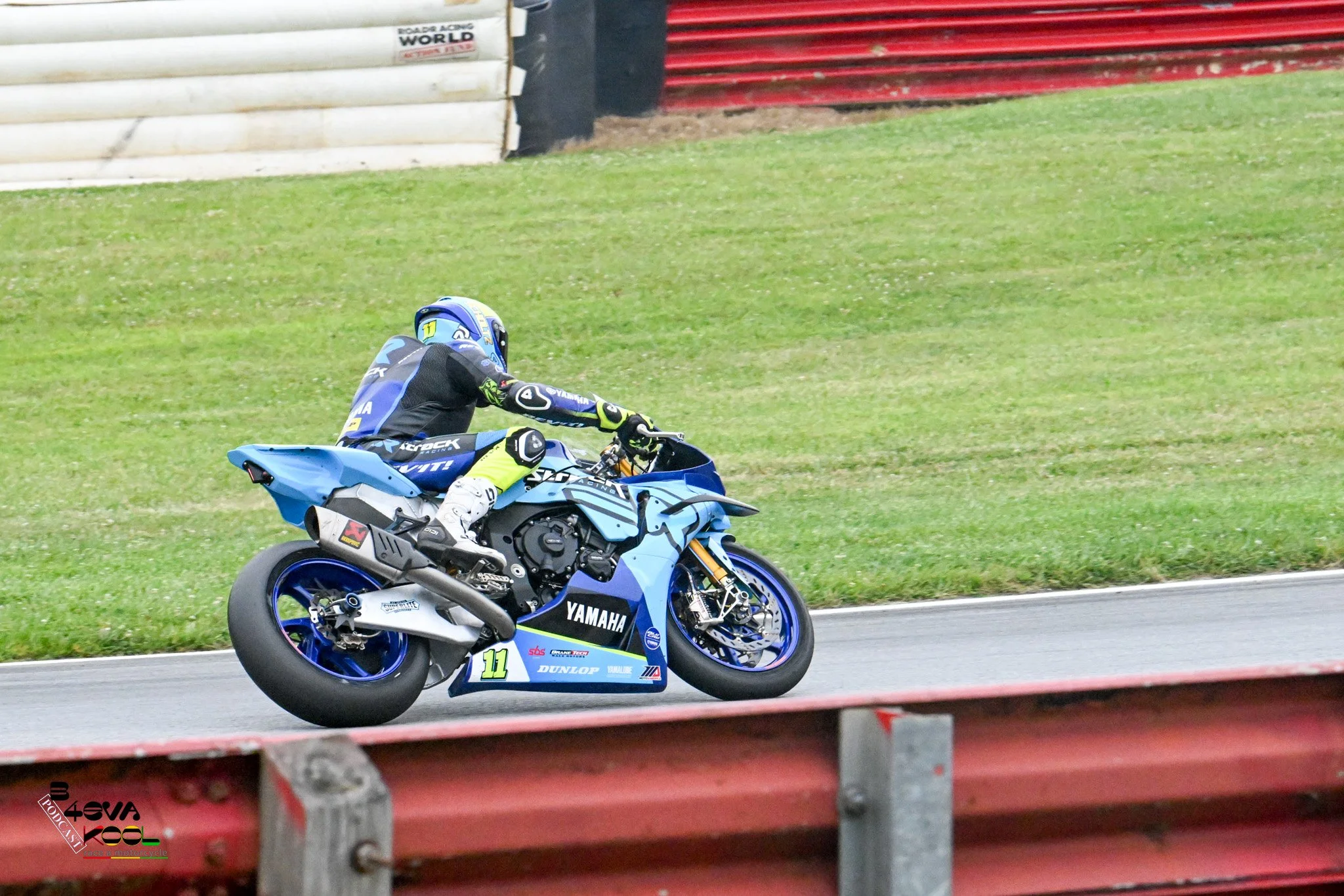 A motorcycle racer wearing a blue helmet and black racing suit riding a blue Yamaha motorcycle on a racetrack, with green grass and red barriers in the background.