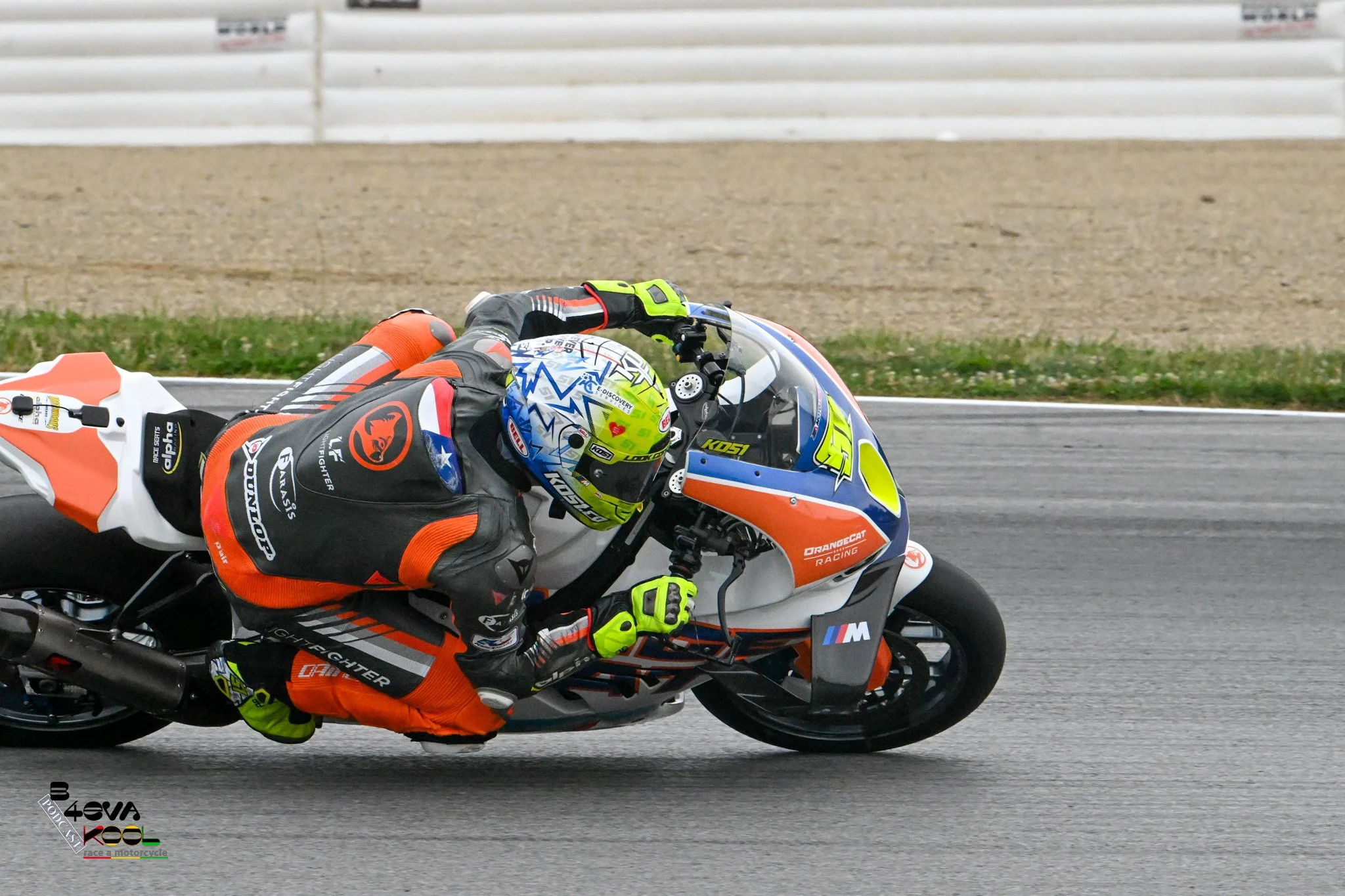 Motorcycle racer leaning into a turn on a race track, wearing colorful racing gear and a helmet, with a gravel and grass background.