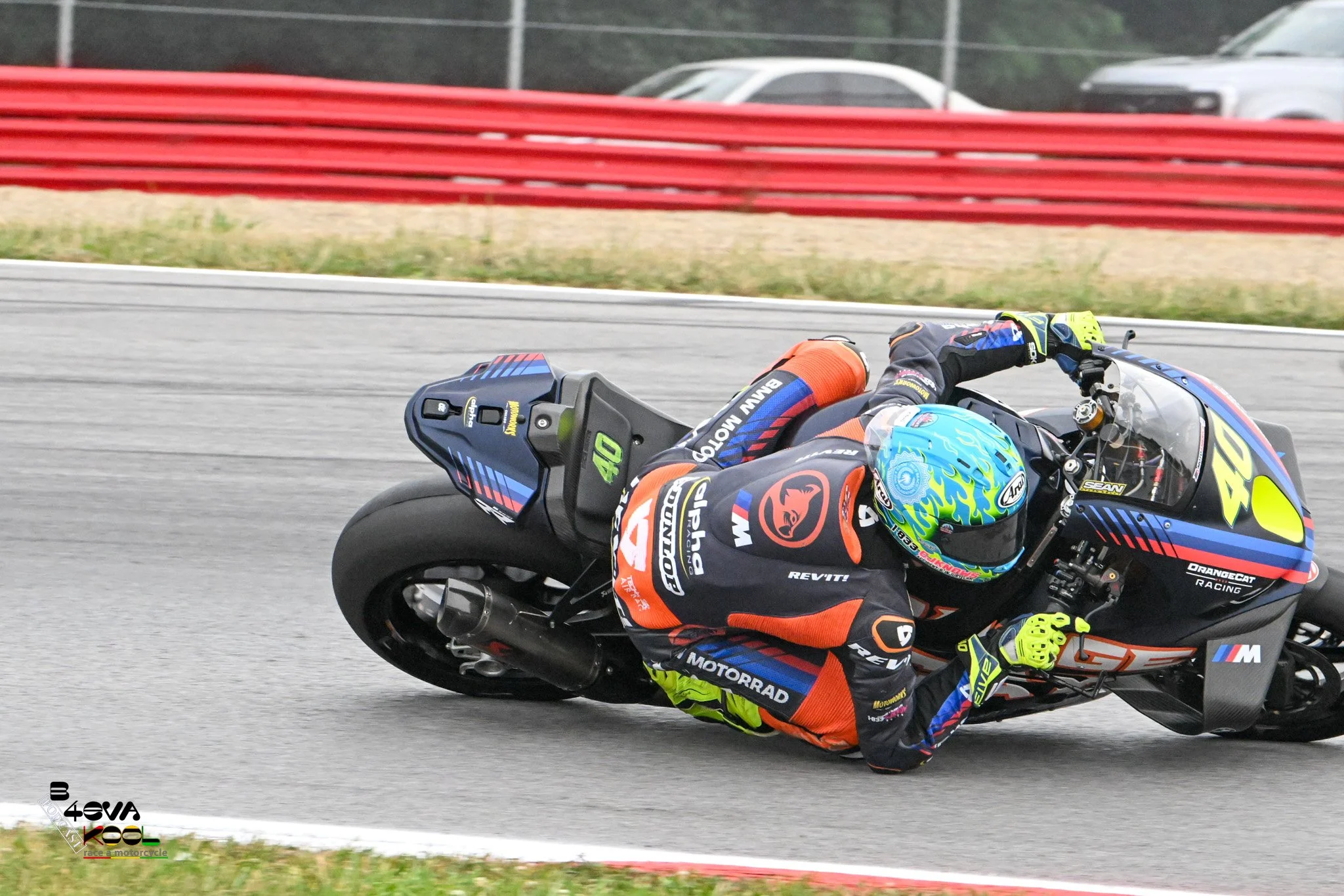 A motorcycle racer leaning closely to the ground while navigating a race track. The rider is wearing a blue and yellow helmet and a black racing suit with colorful accents and sponsor logos. The motorcycle is black with blue and yellow details, and t