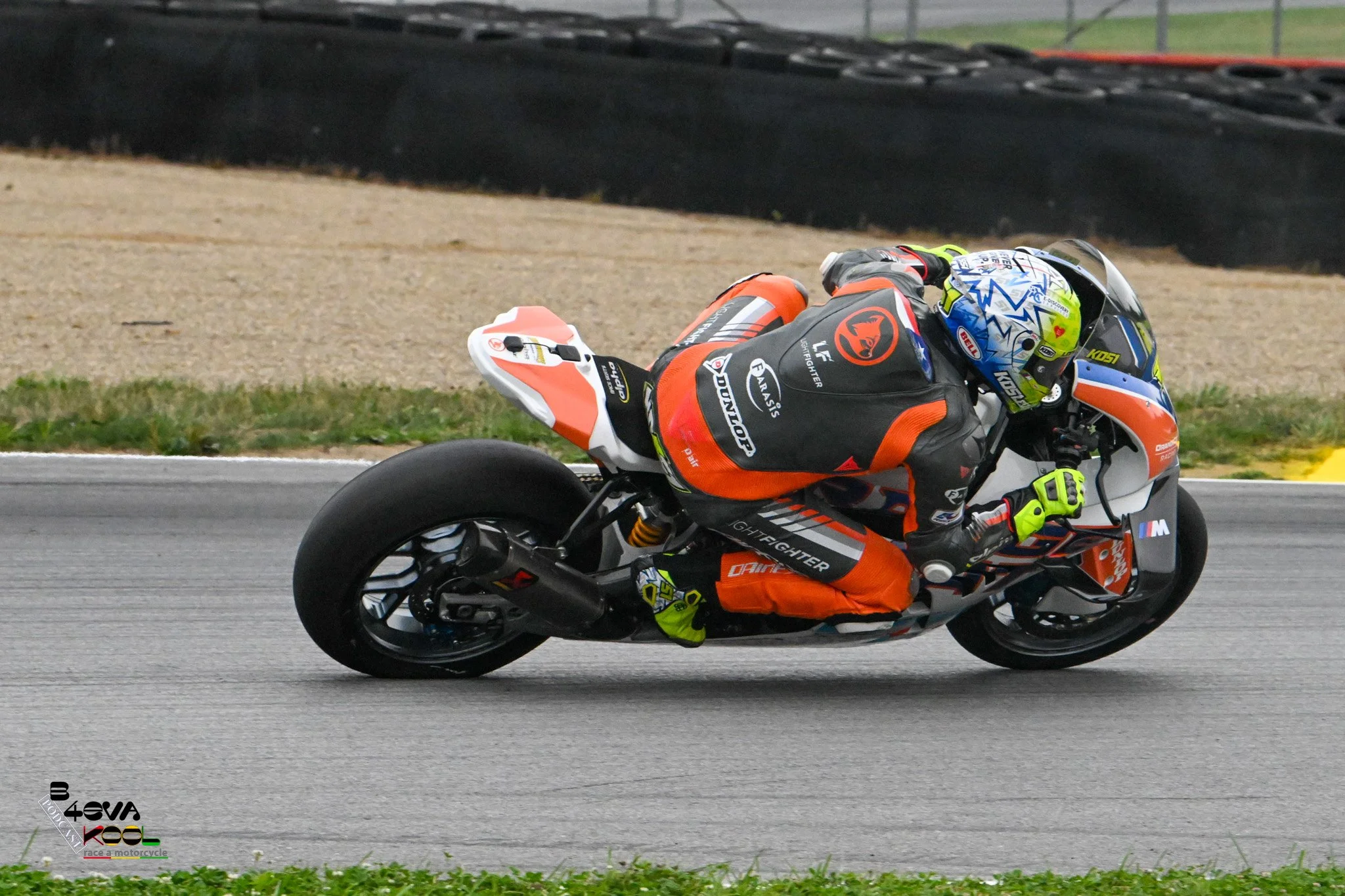A motorcycle racer wearing a colorful helmet and a black and orange racing suit leans into a turn on a racetrack.