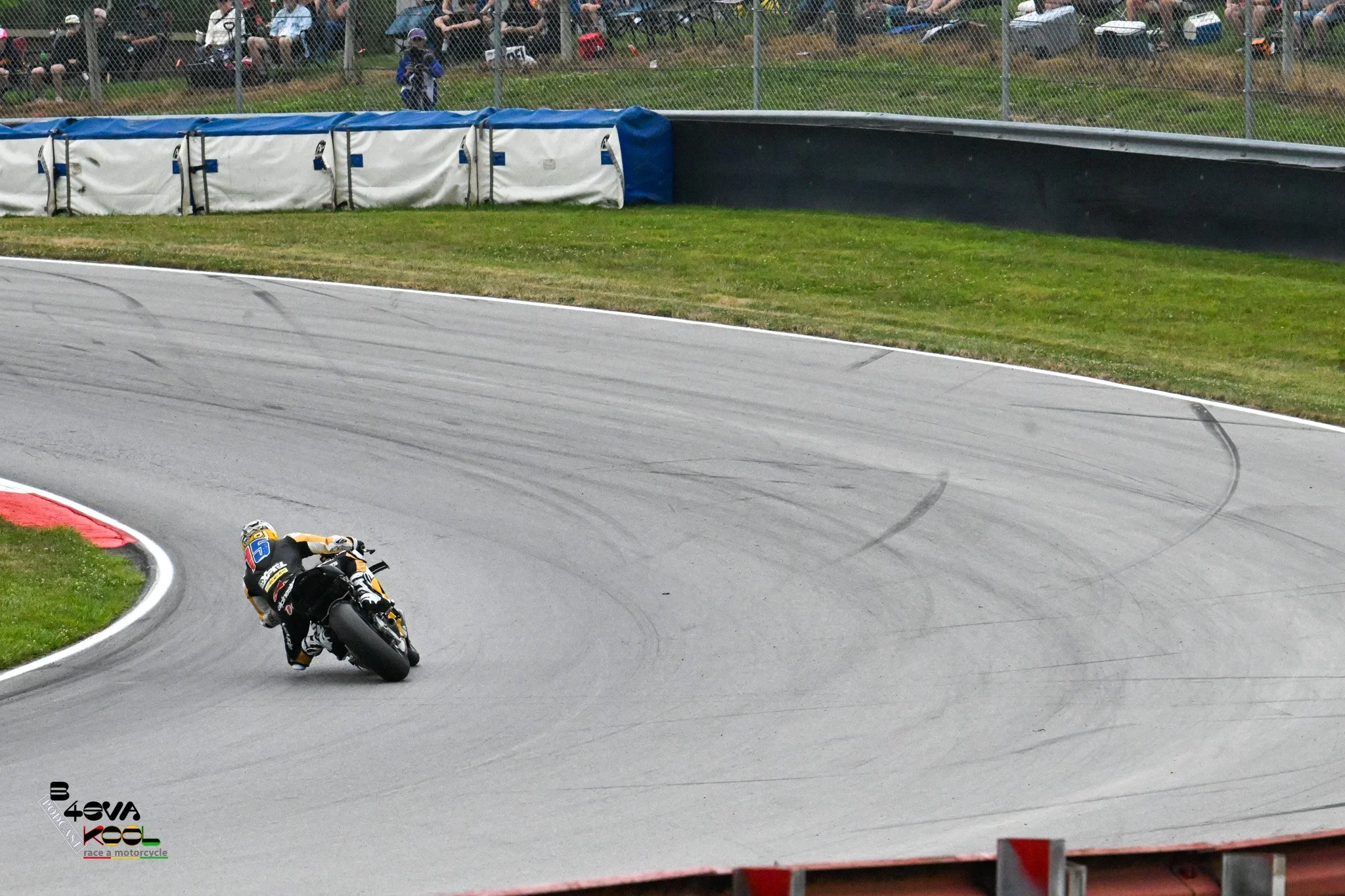 Motorcycle racer leaning sharply into a turn on a racetrack with spectators watching from behind a fence.