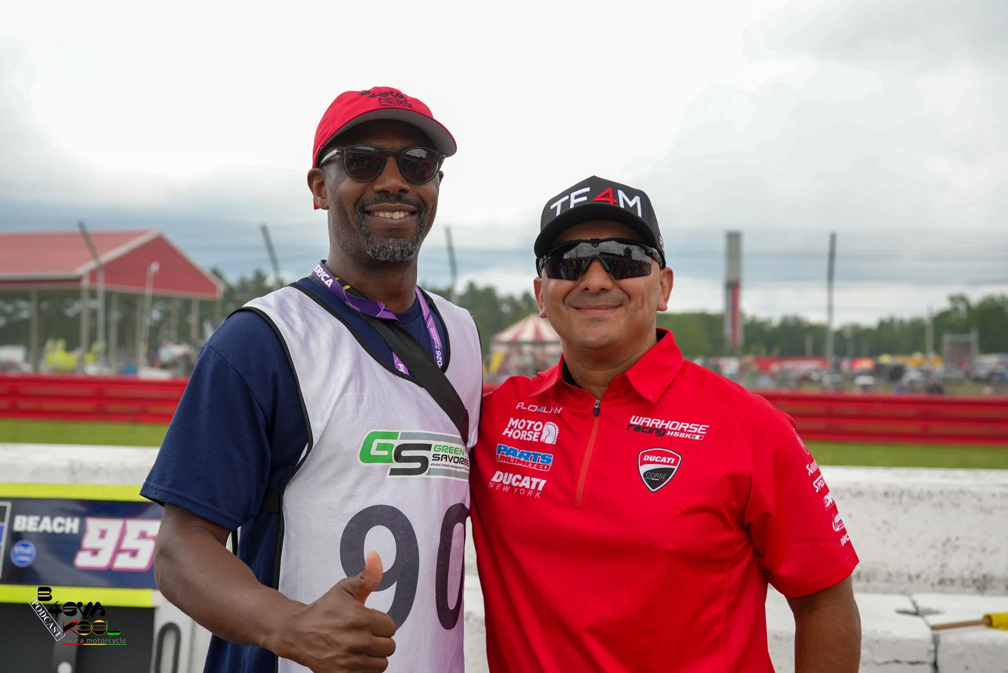 Two men pose together at a racing event, one wearing a white and blue racing shirt with sunglasses and a red cap, the other in a red racing shirt and black cap with sunglasses, smiling.