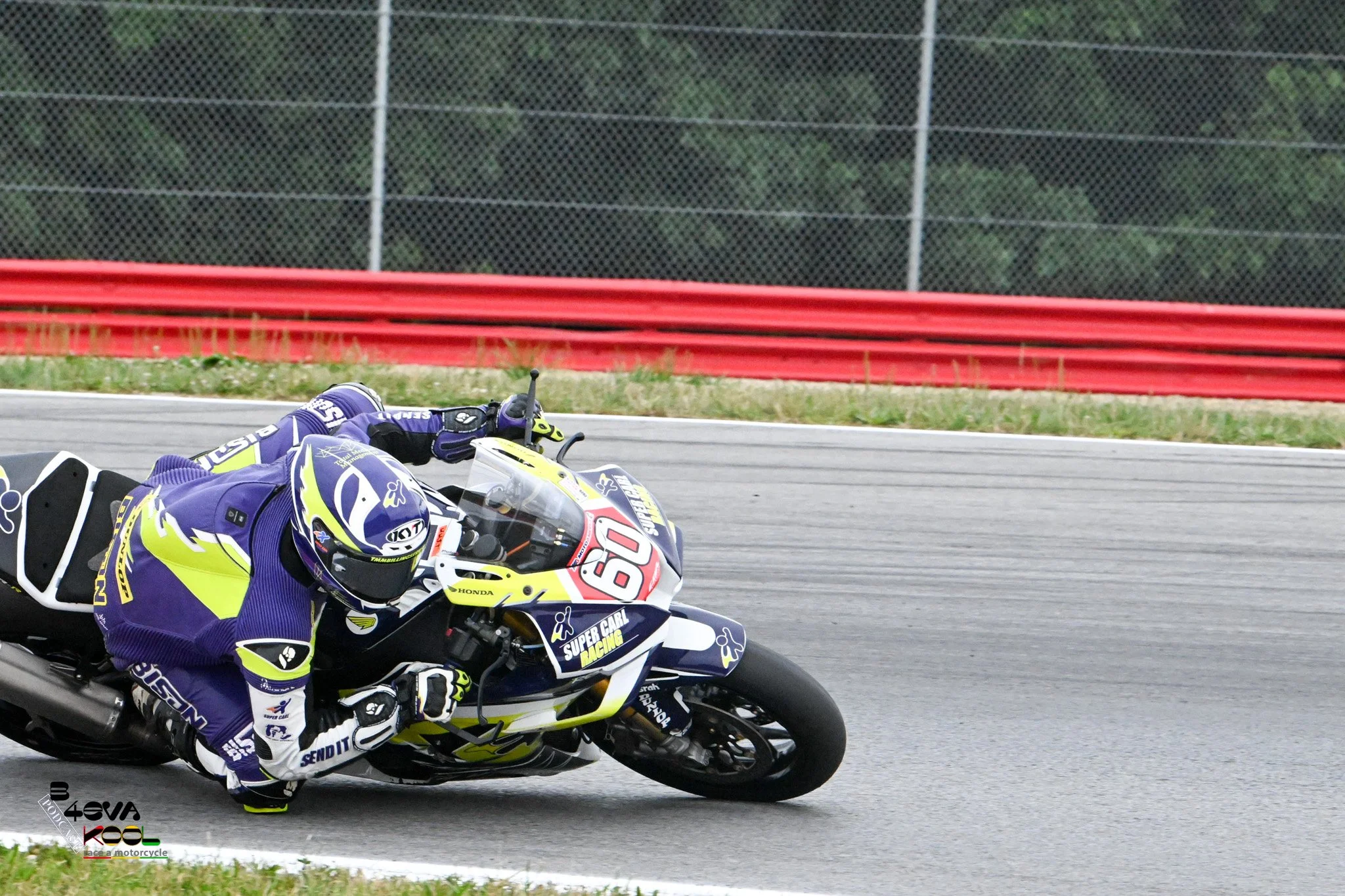 A motorcyclist in a purple and yellow racing suit and helmet leaning into a turn on a racetrack, riding a white motorcycle with racing decals.