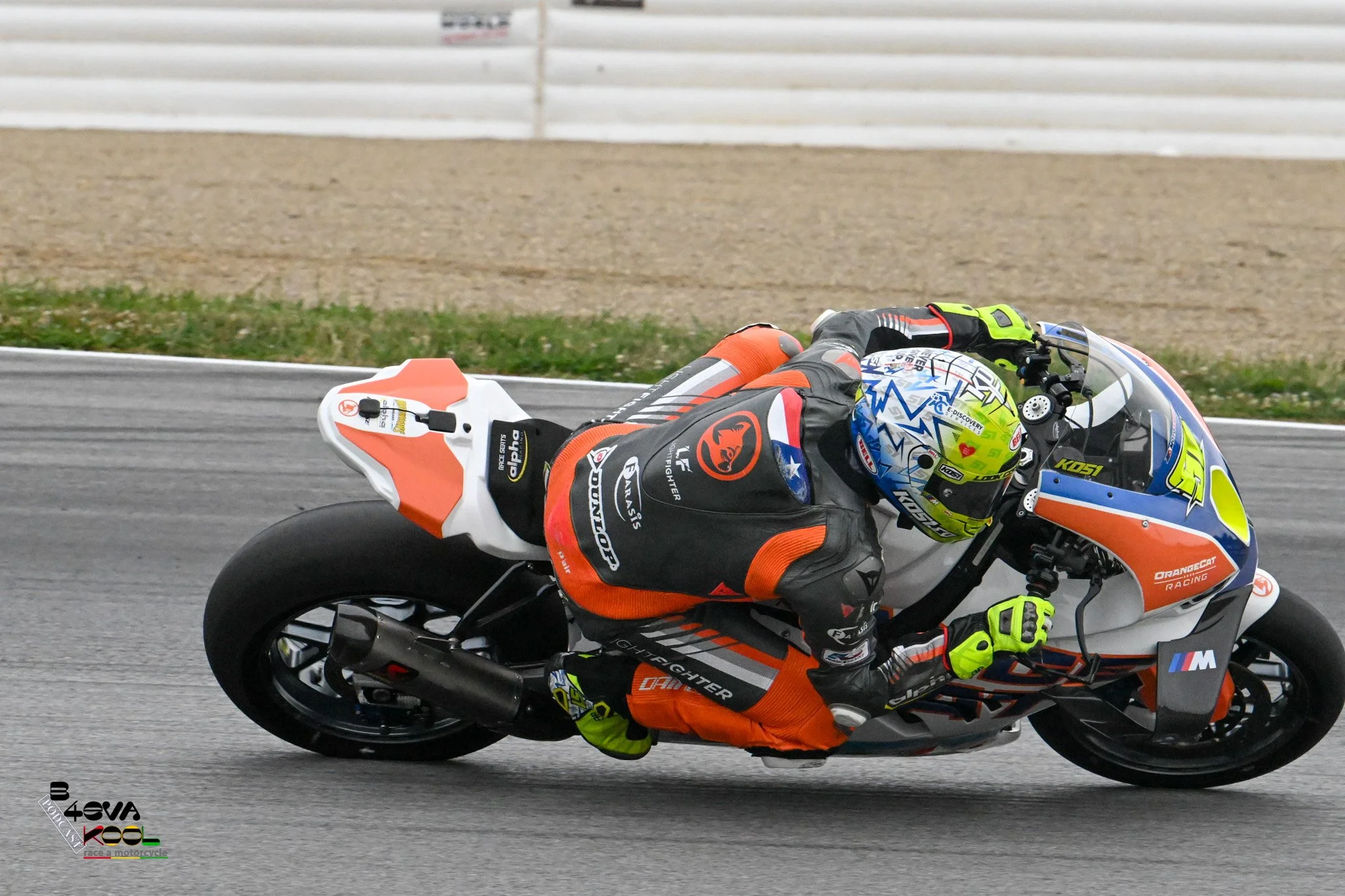Motorcycle racer in orange, black, and blue gear leaning into a turn on a racing track.