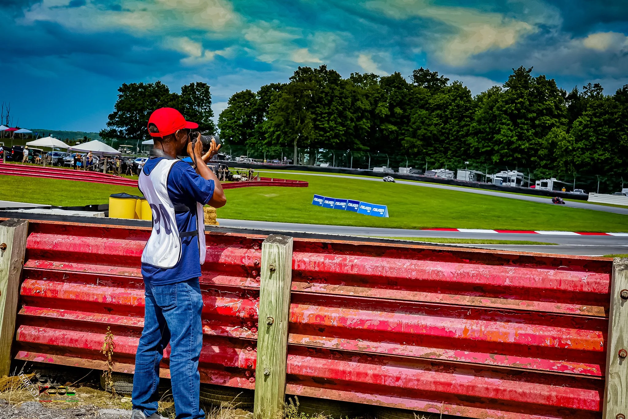 A person wearing a red cap and blue shirt taking a photograph at a race track with trees and spectators in the background.