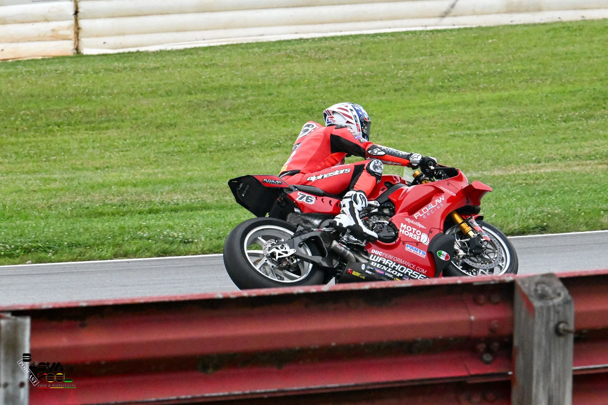 Motorcycle racer in red riding suit leaning into a turn on a race track with grass and a wooden barrier in the background.
