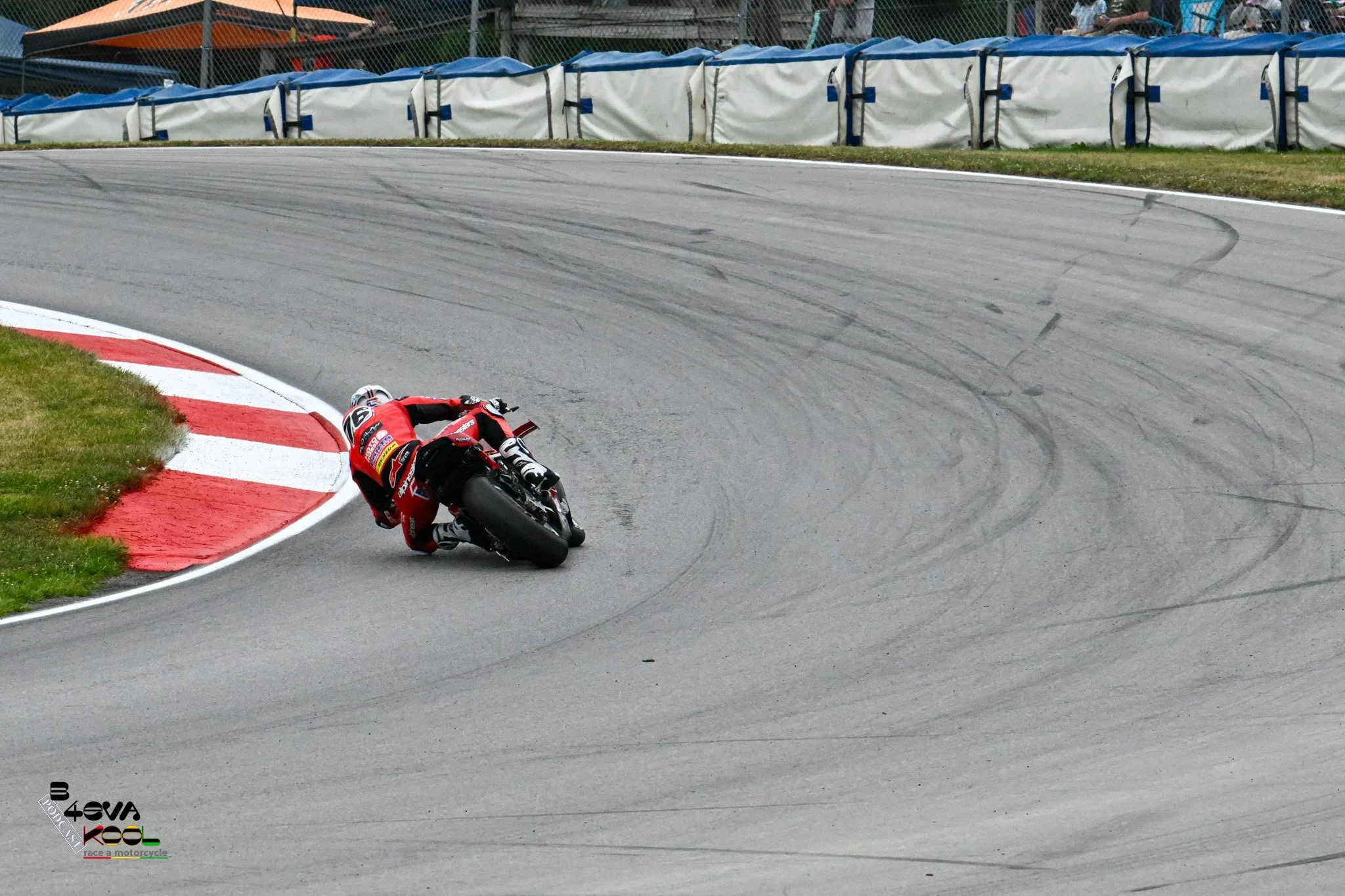 A motorcycle rider in a red racing suit crashes and falls on a racetrack near a curve marked with red and white paint, with tire marks visible on the asphalt.