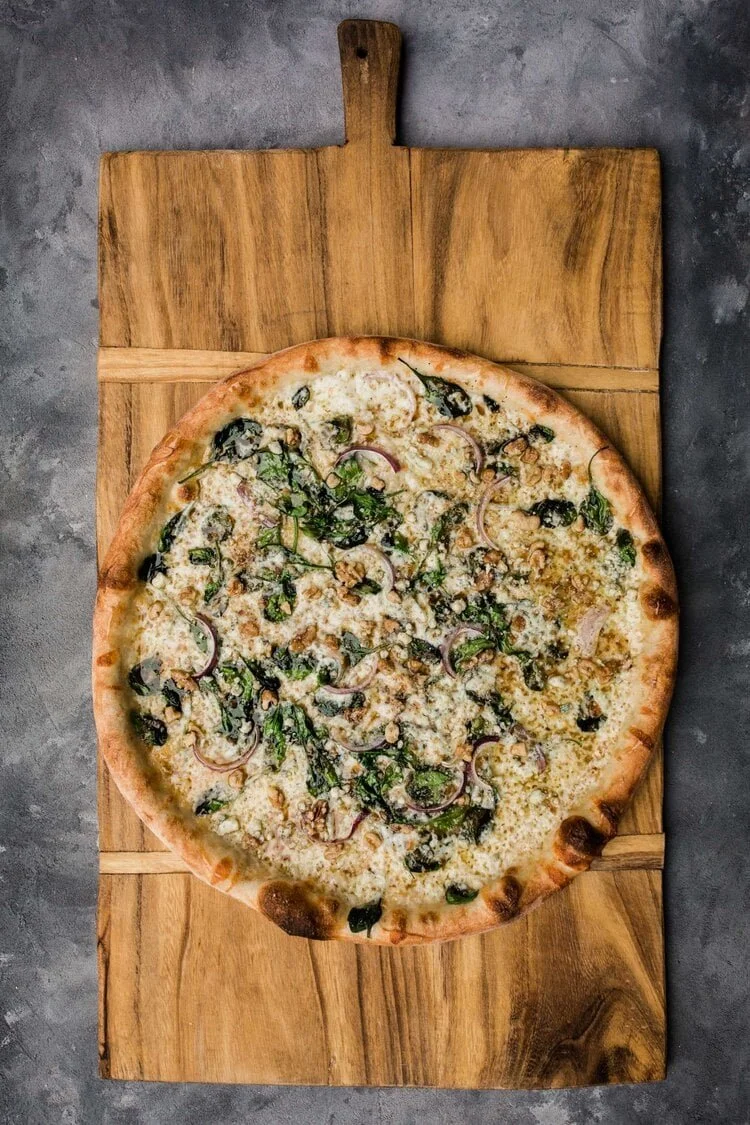 A cheese pizza topped with herbs and red onions, pesto and walnuts on a wooden cutting board.