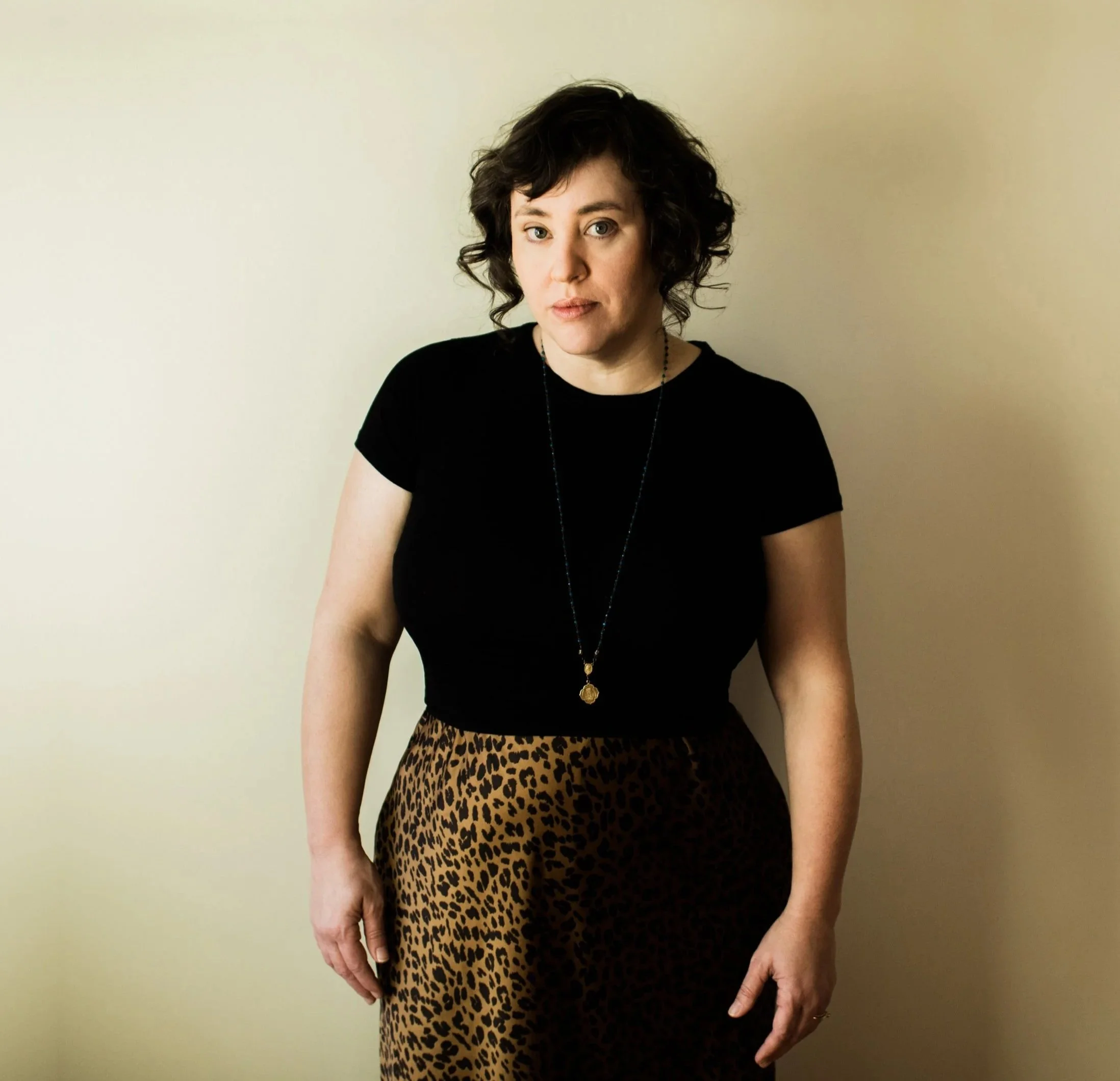 Asheville photographer, Jennifer Callahan, wearing a black shirt and print skirt, standing against a plain beige wall.
