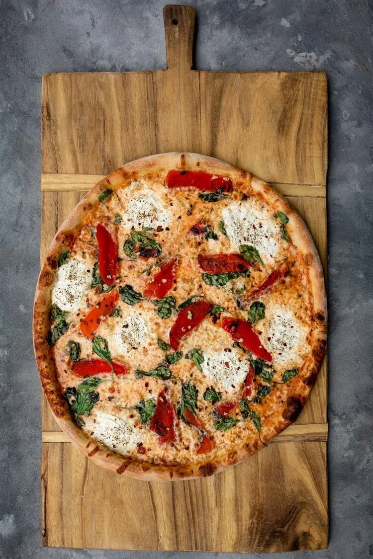 Pizza with roasted red bed pepper, basil leaves, mozzarella cheese, and red pepper flakes on a wooden pizza peel.