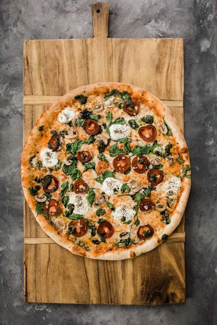 Pizza on a wooden serving board, topped with bresh basil, roasted garlic , tomatoes slices, and fresh Burrata.