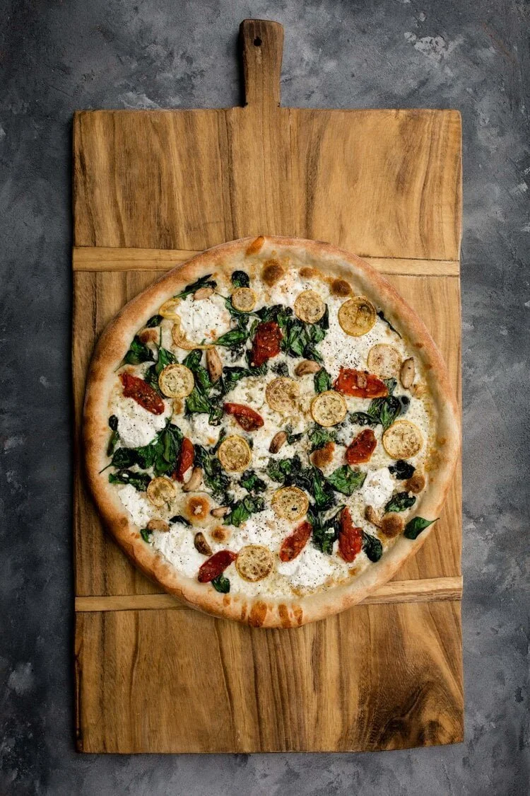 Pizza on a wooden serving board, topped with spinach, roasted garlic slices, sun-dried tomatoes, and yellow squash.