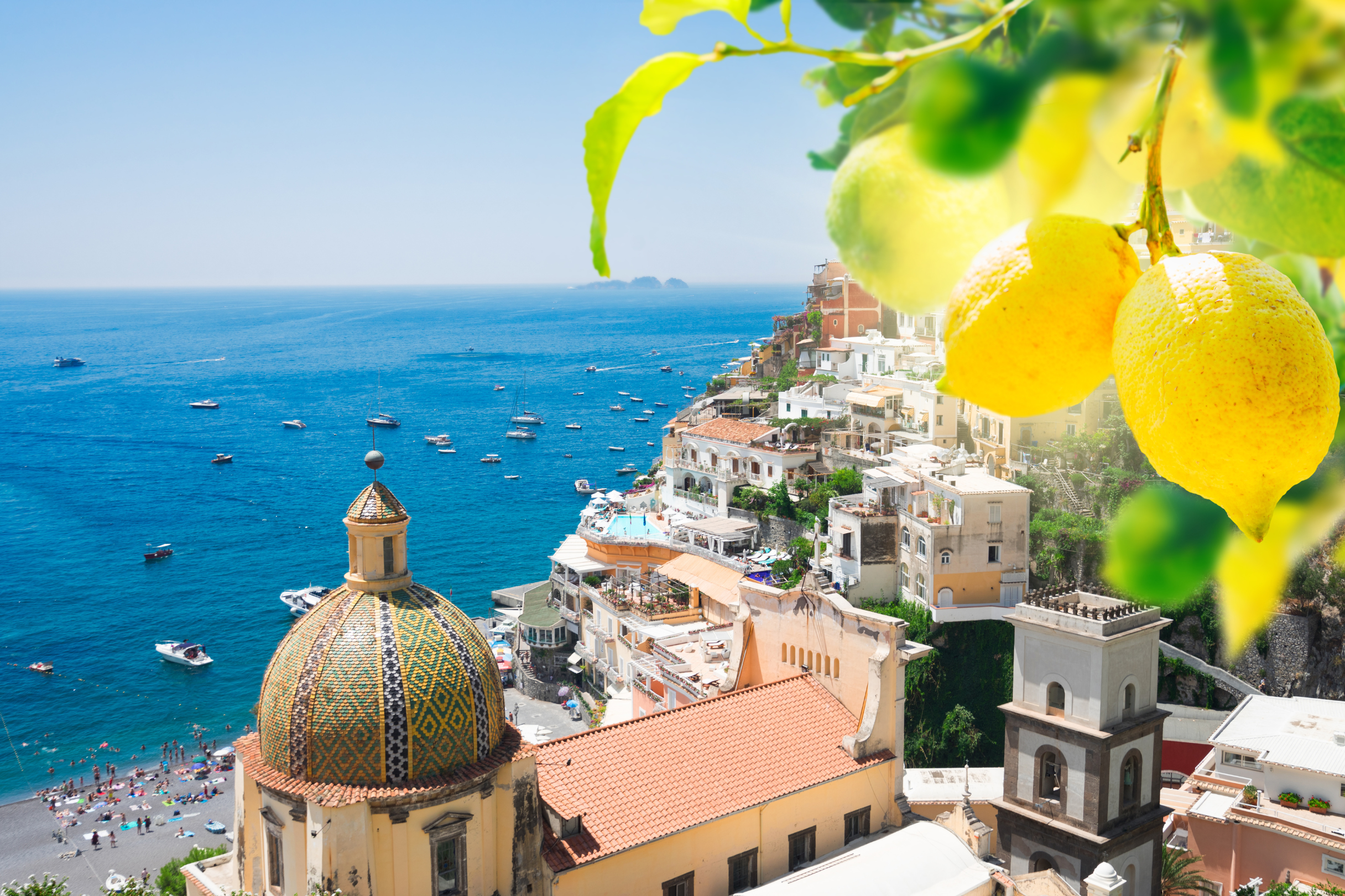 View of the Amalfi Coast with lemons in the foreground, colorful buildings, a church dome, and boats on the Mediterranean Sea.