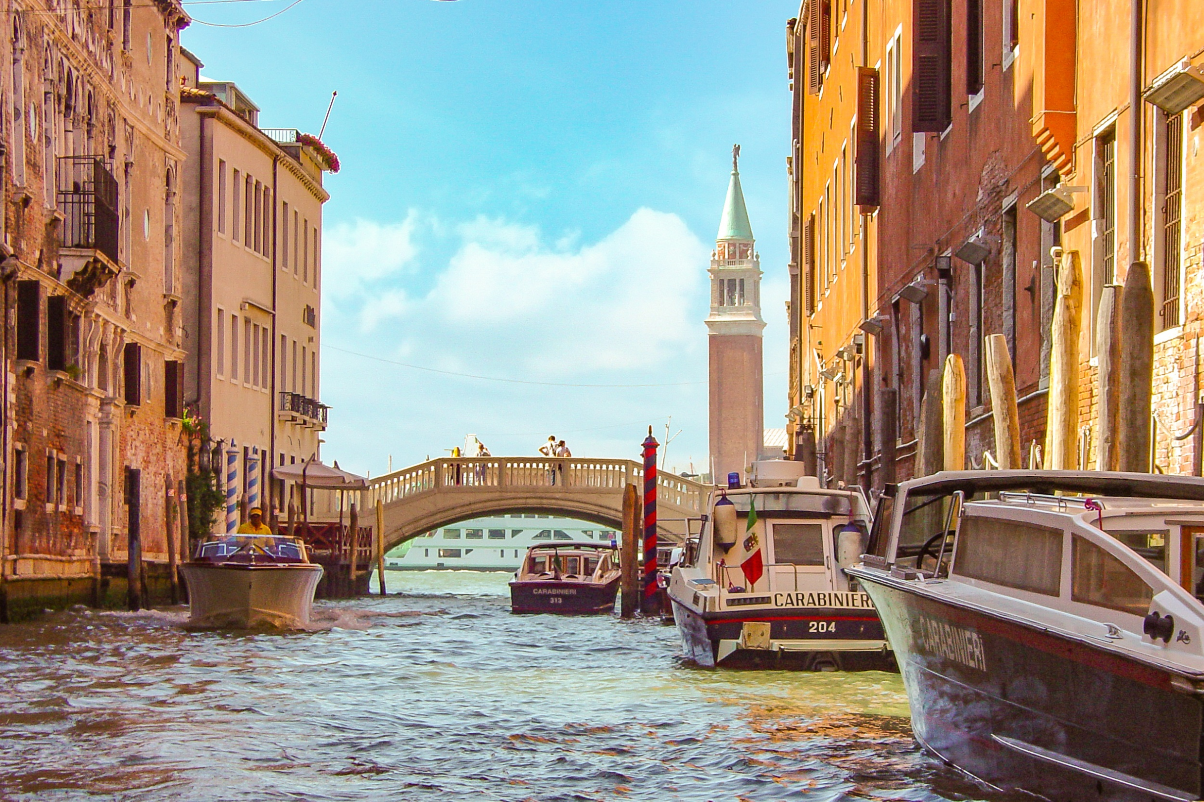 Venice canal scene with gondolas, bridge, and bell tower on a sunny day.