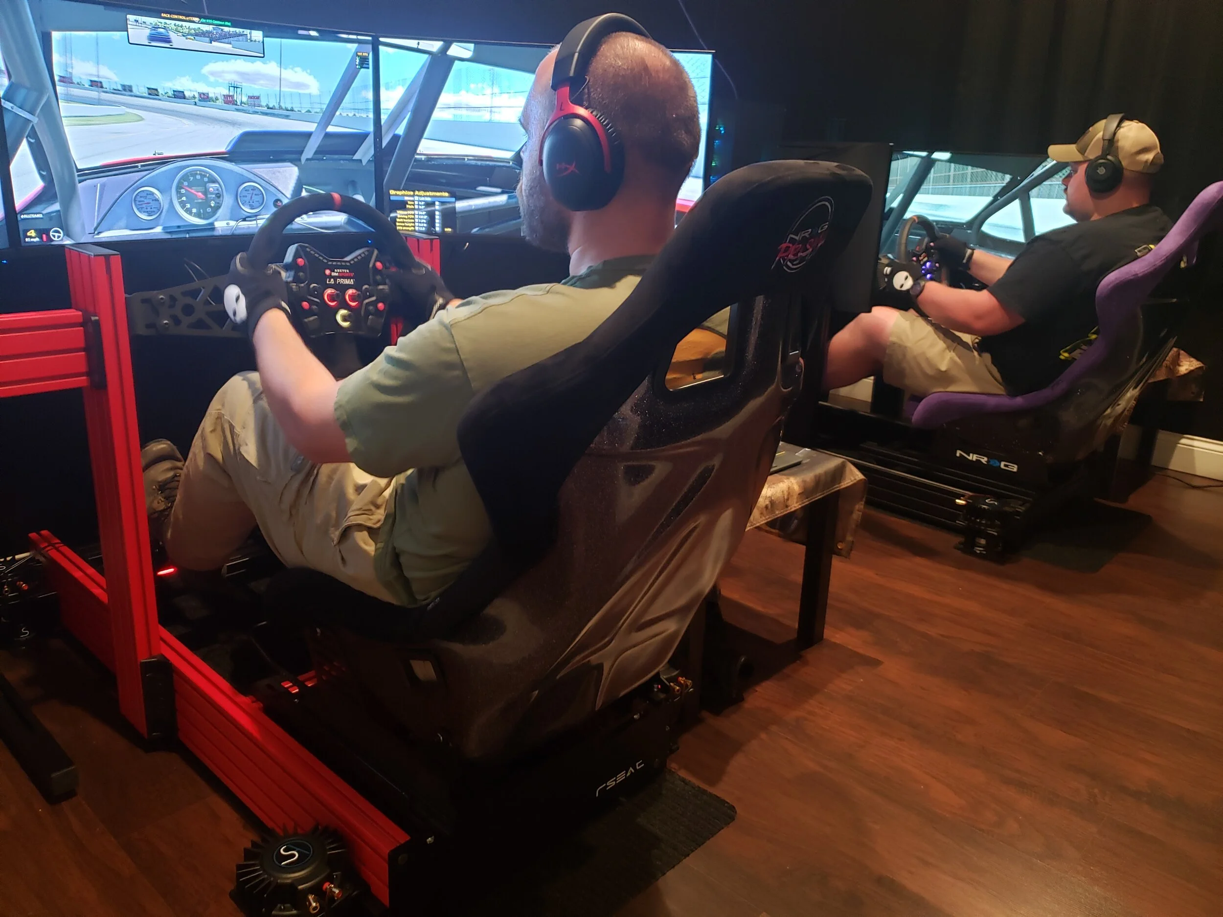 Two men in a racing simulator setup, each sitting in a racing seat with a steering wheel, pedals, and multiple screens displaying a racing game or simulation.