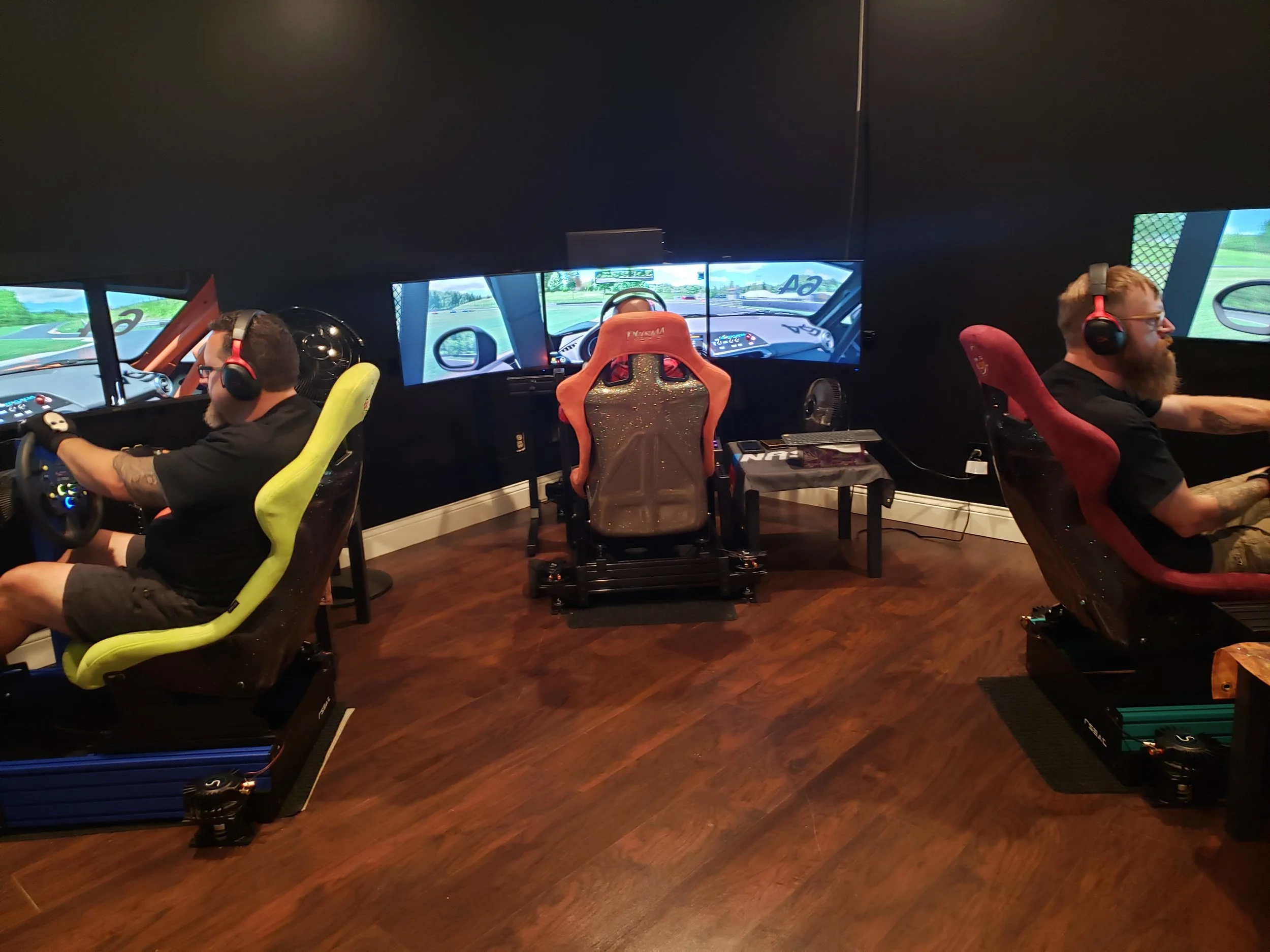 Two men wearing headphones sitting in racing seat simulators, each facing multiple screens showing a racing game, in a room with wooden floor and black walls.