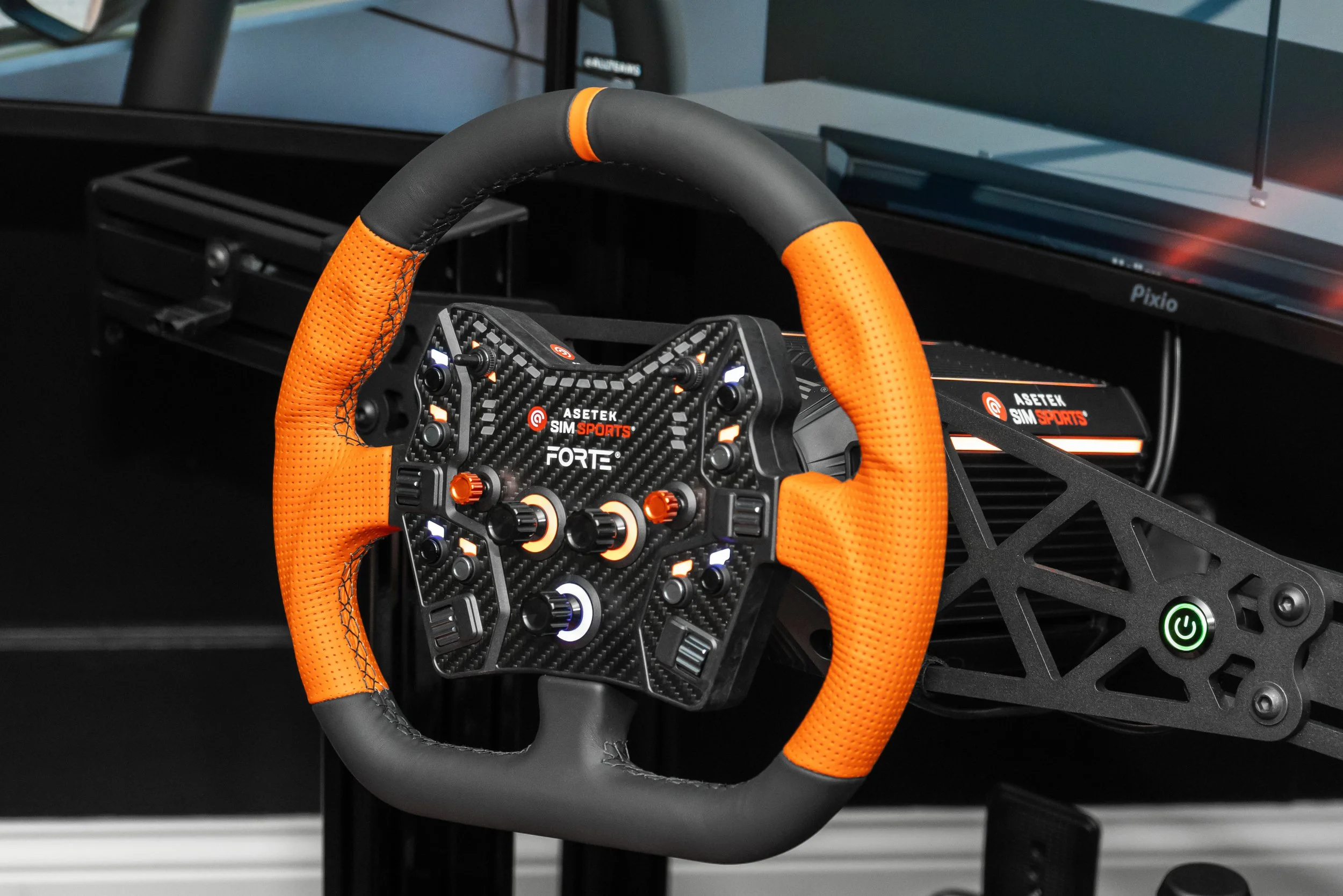 Racing simulator steering wheel with orange and black grip, mounted on a black frame, with a green power button, and a monitor in the background.