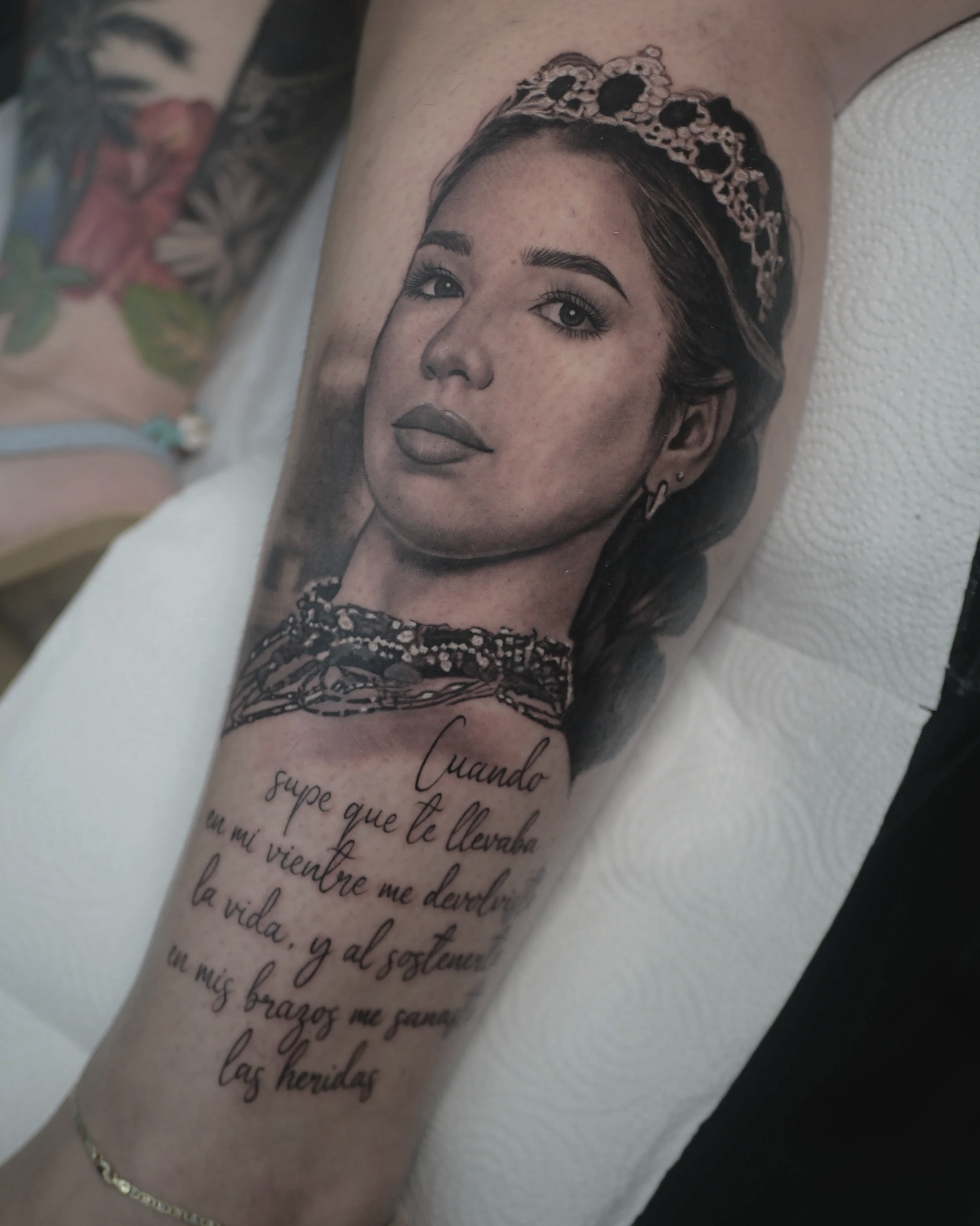 Tatuaje en el brazo de una mujer con una corona, acompañado de una cita en español que dice: 'Cuando supe que te llevaba en mi vientre me derrumbé la vida, y al sostenerte un mess brazos me sanaron las heridas'.