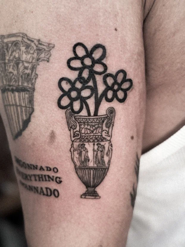 Tatuaje de un jarrón con flores en forma de corazón, rodeado por figuras humanas y texto en inglés.