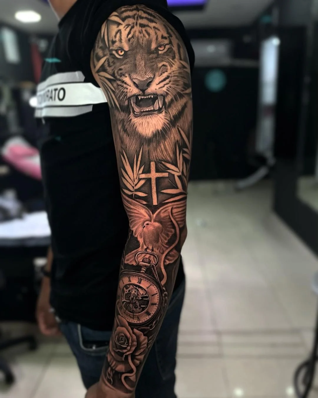 Tatuaje en el brazo de una persona que muestra un león rugiendo, una cruz, una paloma, un reloj de bolsillo y varias rosas en estilo gráfico en tonos negros y grises.