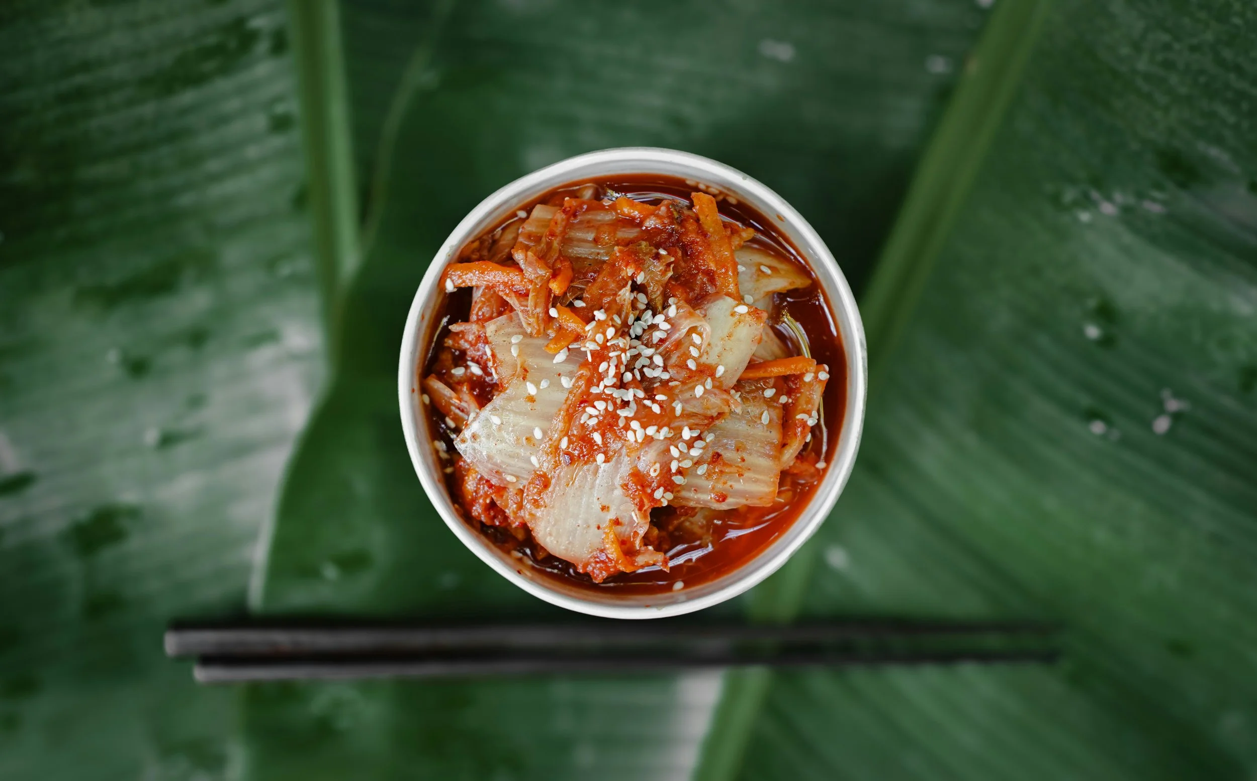 A vibrant close-up of a bowl of kimchi, served with chili paste and sesame in a bowl, in front  of a background of palm leaves. The image showcases one of the most popular types of fermented foods from Korea.