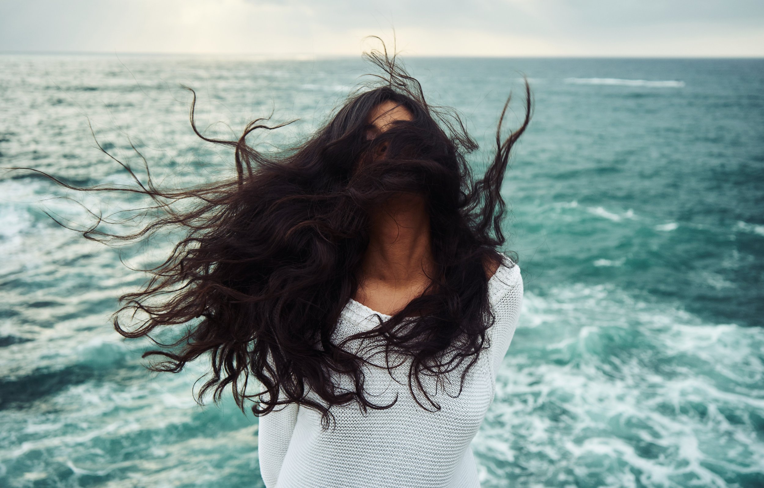 Woman with windblown natural hair by the ocean, showcasing effortless texture and a natural hair routine inspired, air-dried look.