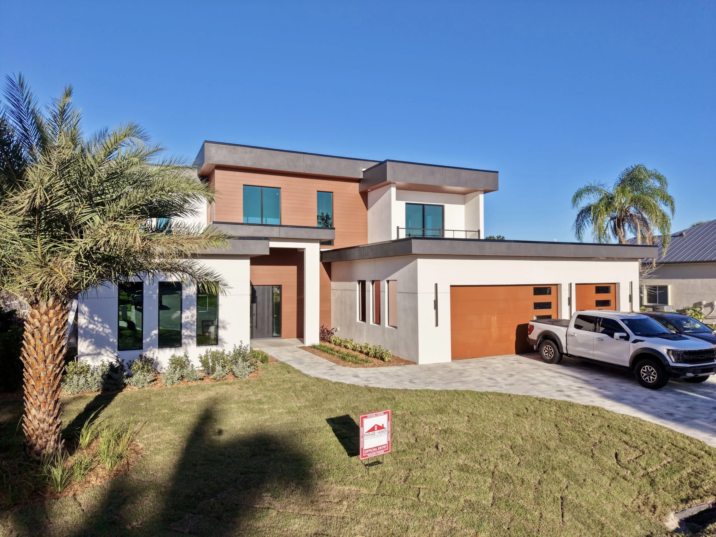 Modern two-story house with large windows, a garage with an orange door, and a driveway. There is a green lawn, palm trees, and a small sign that says 'Official Entry' in the front yard. The sky is clear and blue.