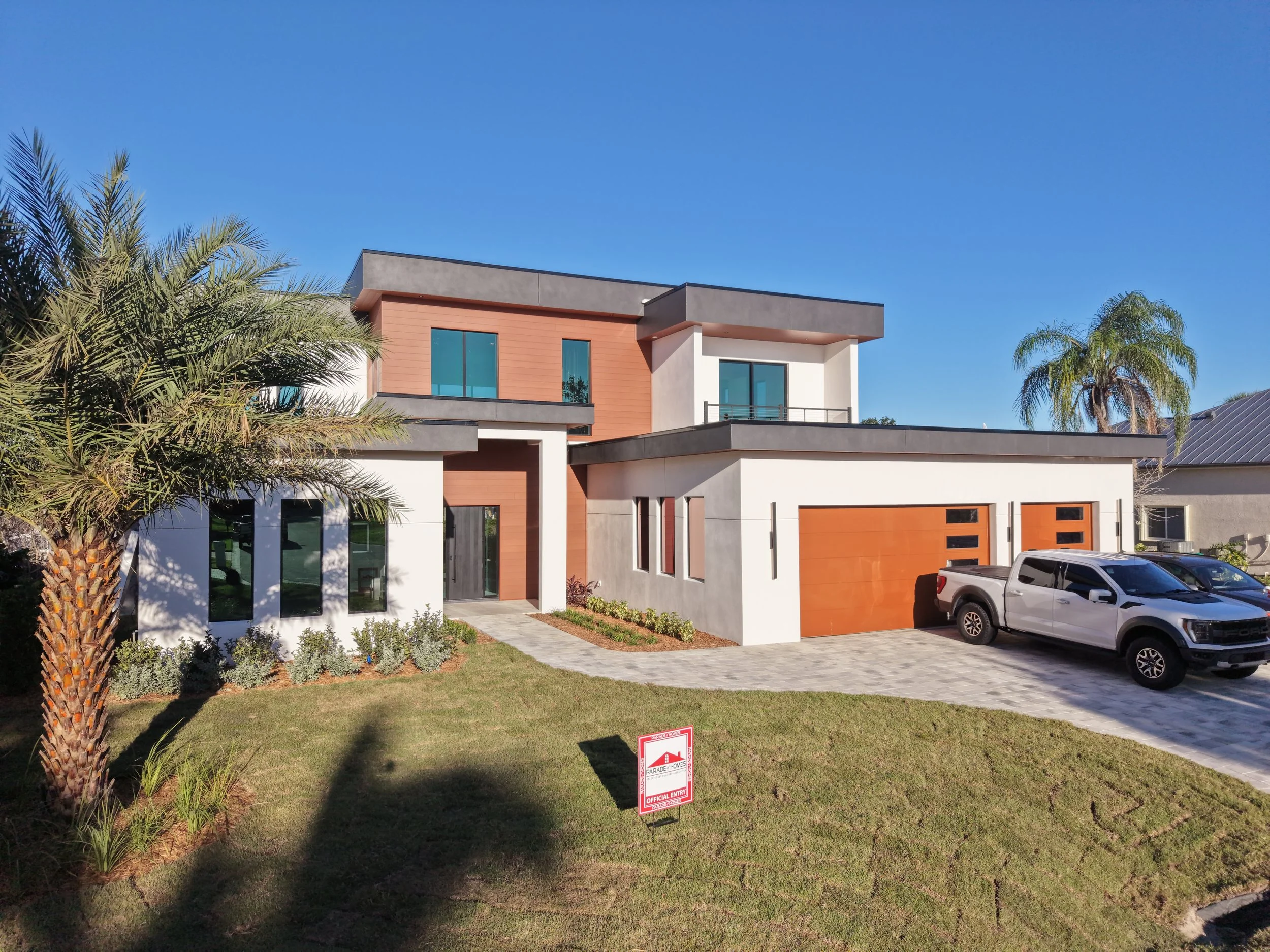Modern two-story house with large windows, a garage with orange doors, and a small front yard with a sidewalk and parked cars. Tall palm trees are visible on the sides, and there is a clear blue sky.