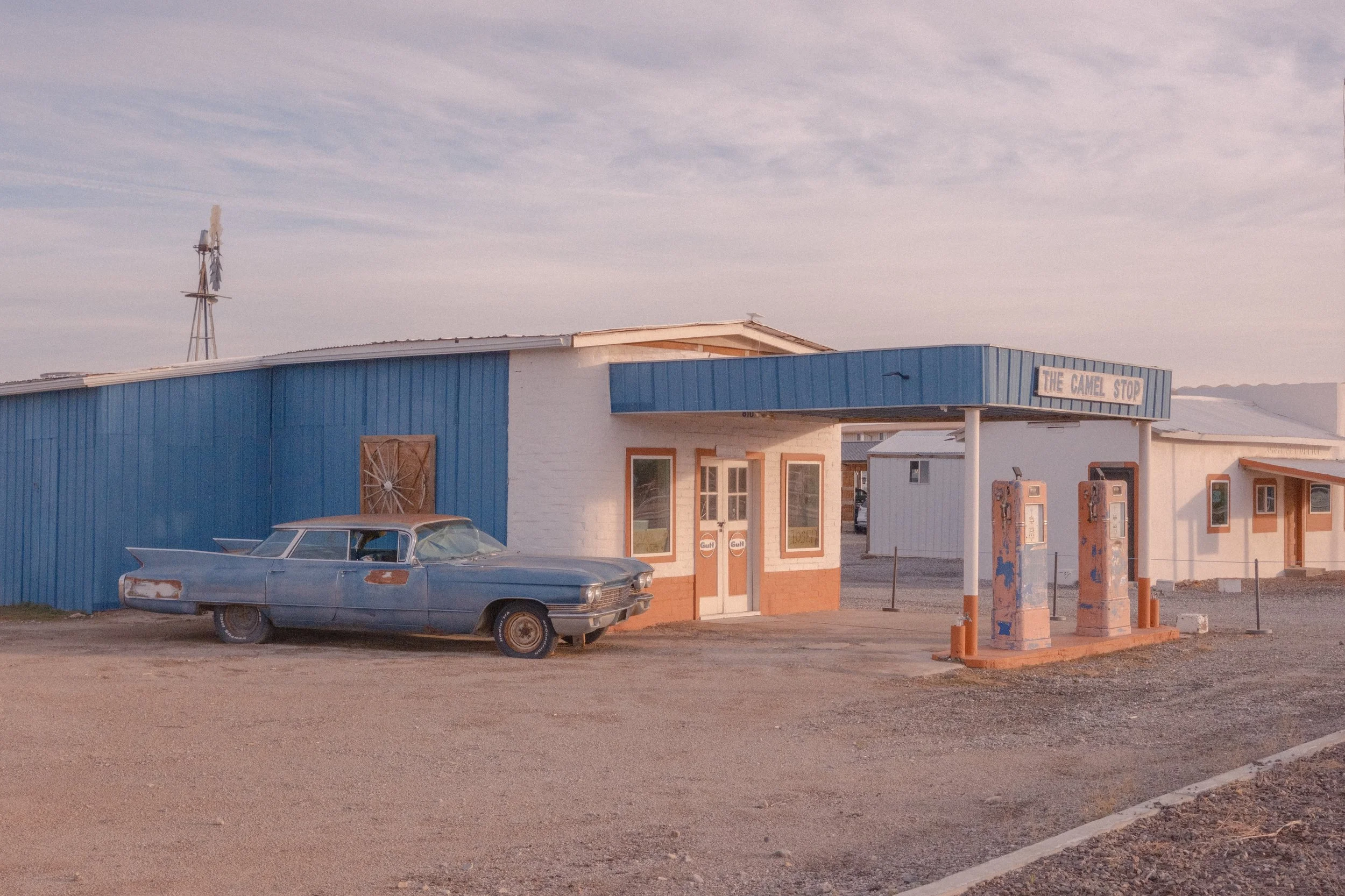 An old car parked in front of a building with a sign that reads "The Camel Stop." The building has a blue exterior with white brick accents, and there are vintage-style automatic teller machines outside. The sky is partly cloudy, and the overall scene suggests a small roadside stop or diner.