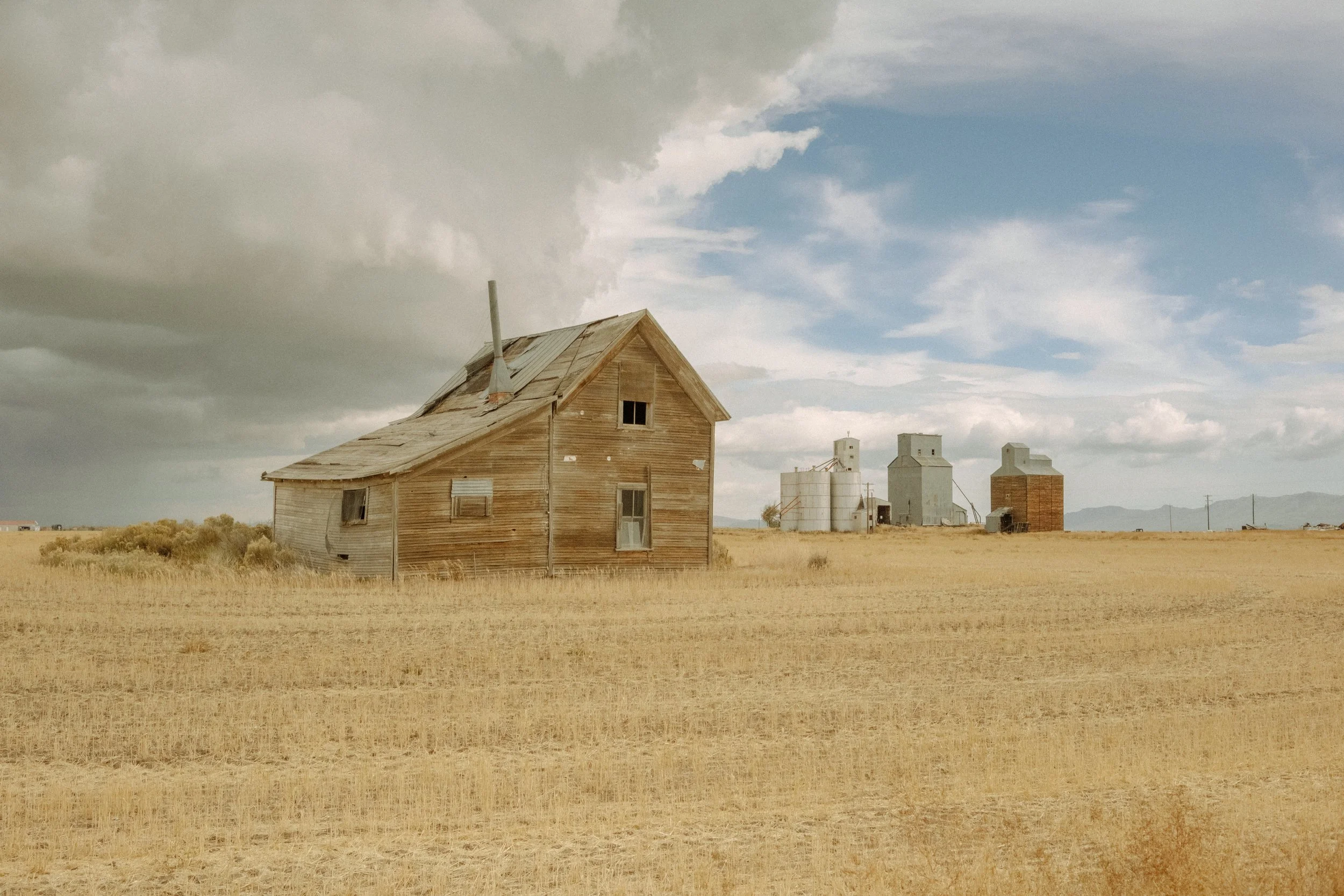 A weathered old wooden house in a wide open field under a partly cloudy sky with large farm silos in the background.