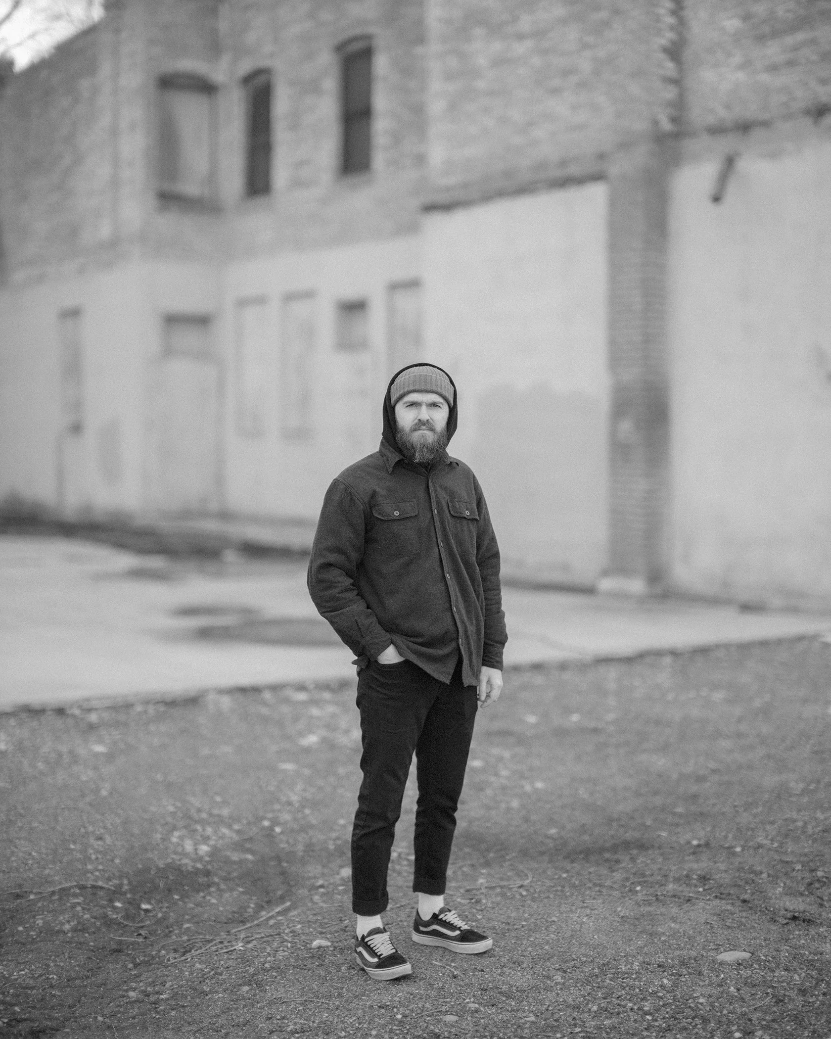 A man with a beard, wearing a hoodie, jacket, and sneakers, stands outdoors on a gravel area in front of a building with brick and plaster walls and tall, narrow windows.