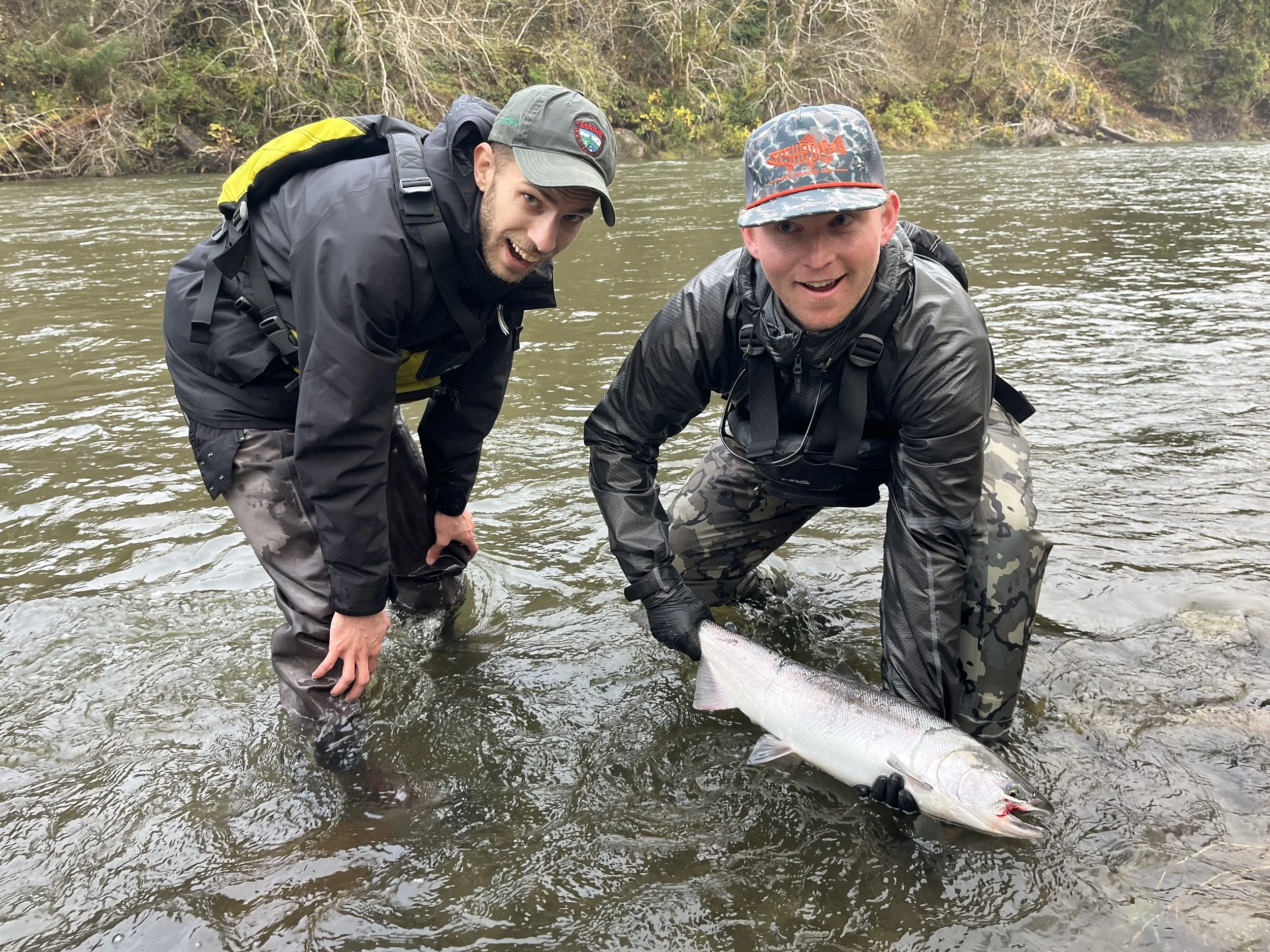 Two men in waterproof clothing are in a river, holding a large fish they caught. They are smiling and looking at the camera. One man is on the left wearing a cap and a yellow backpack, and the other is on the right wearing a camouflage cap and gloves.