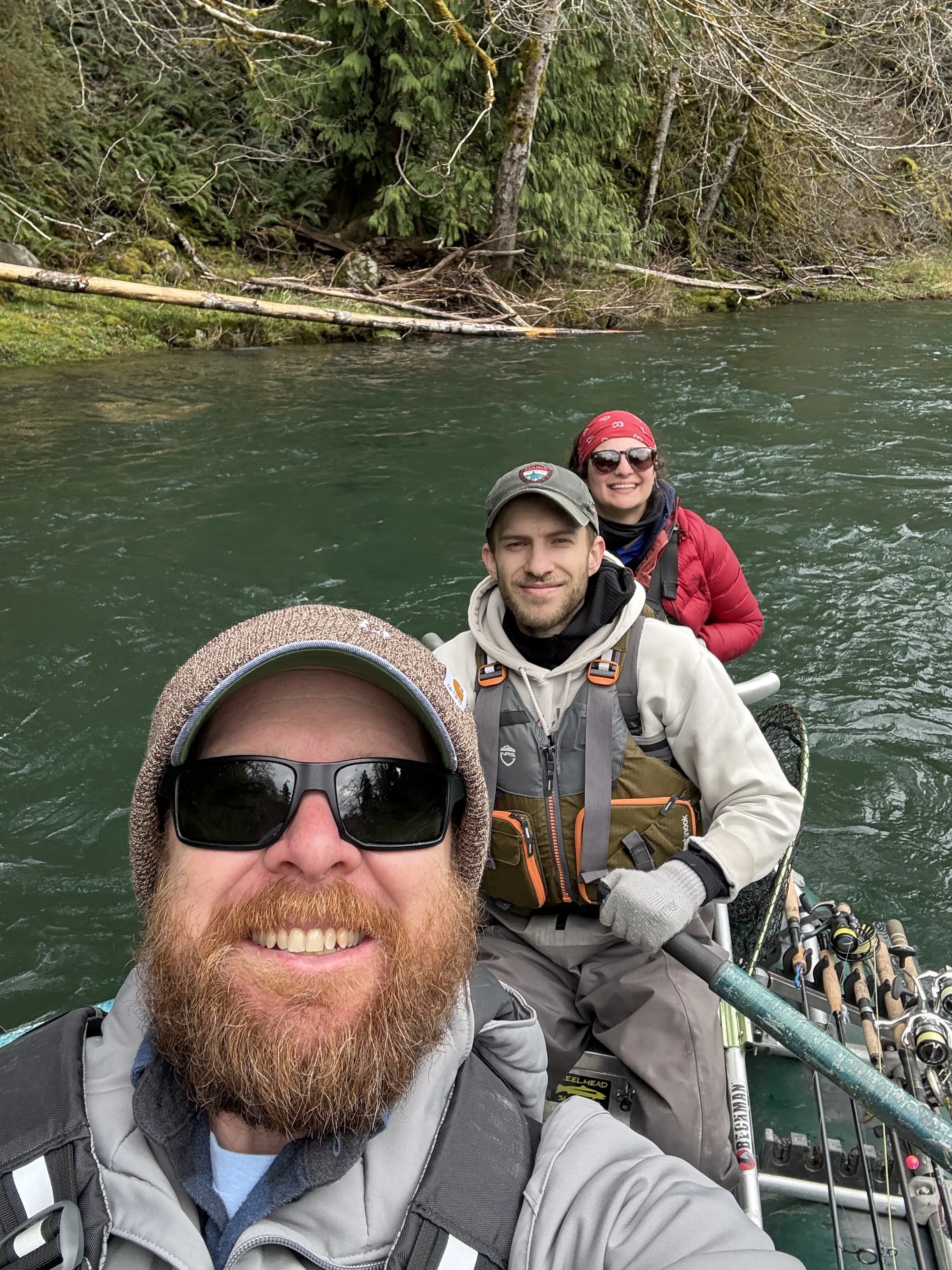 Three people on a fishing boat in a river, surrounded by trees, taking a selfie and smiling.