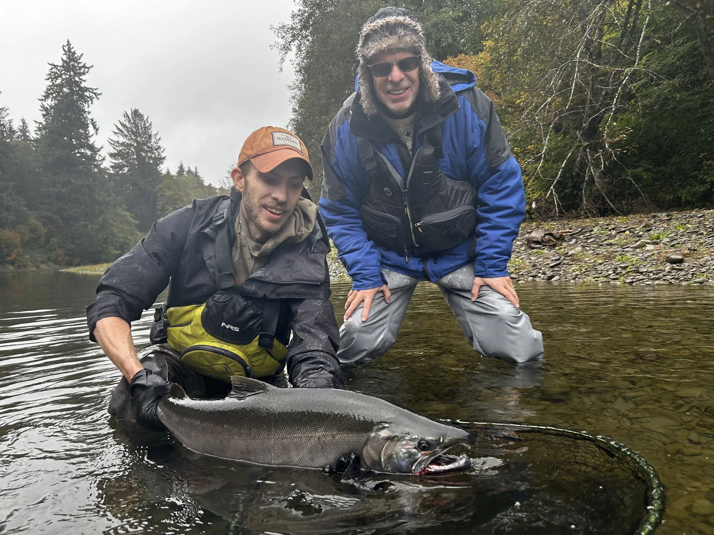 Two men in outdoor gear stand in a river, holding a large Coho Salmon they caught, with a forested landscape and cloudy sky in the background.
